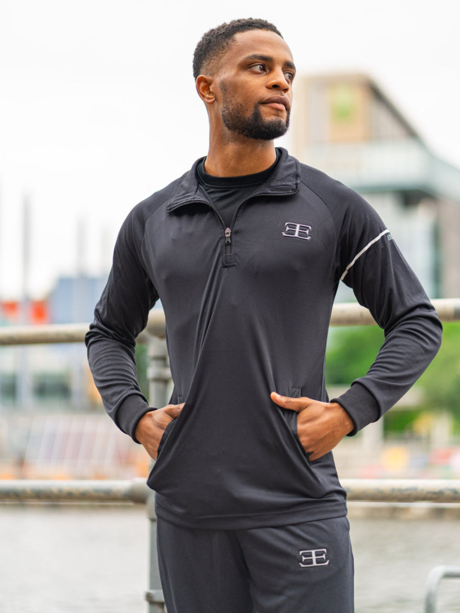 Man wearing a black zip top and black joggers, outside, with Elegante Logo on chest