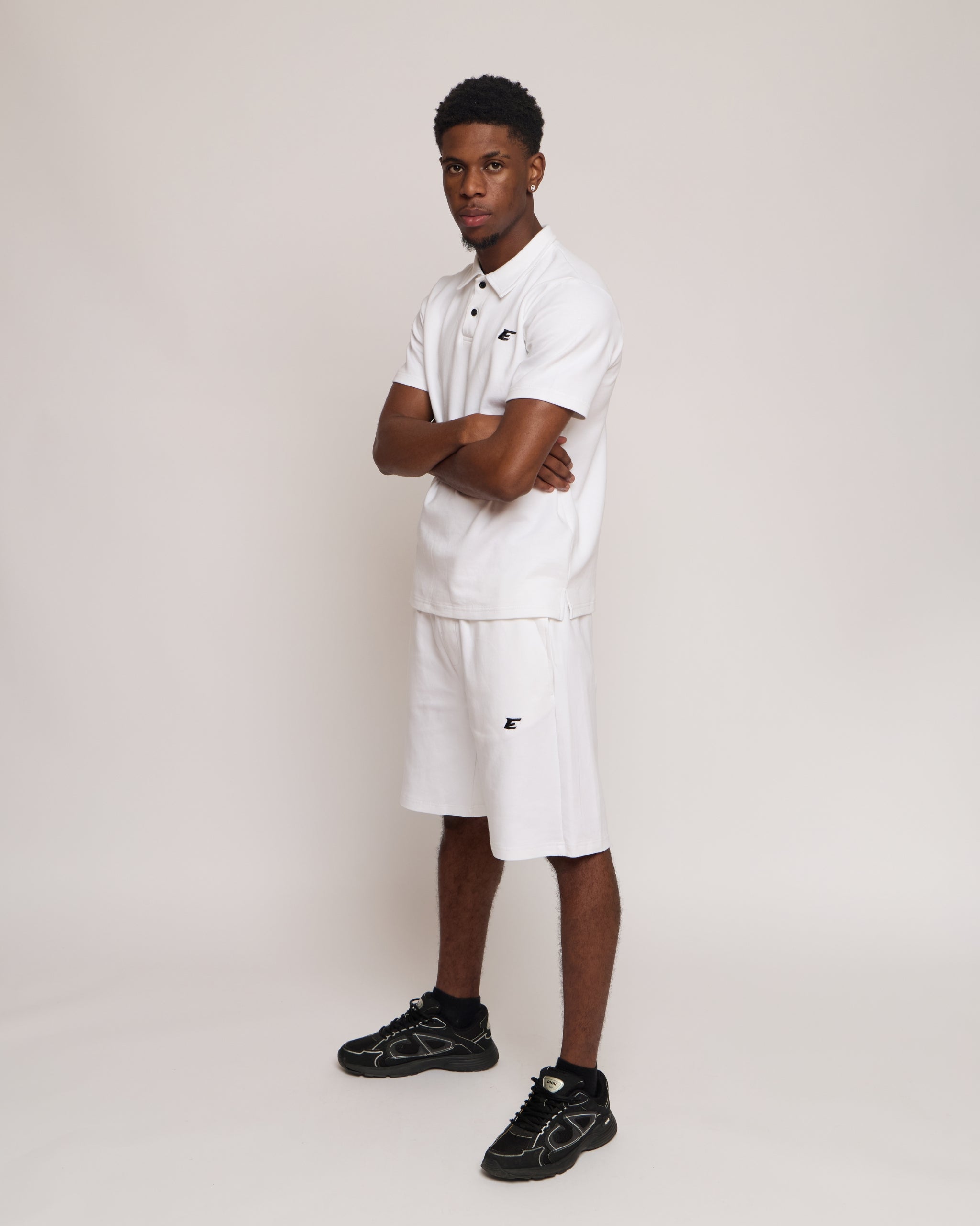 Man wearing a white polo shirt and shorts with an 'E' logo, standing against a plain background