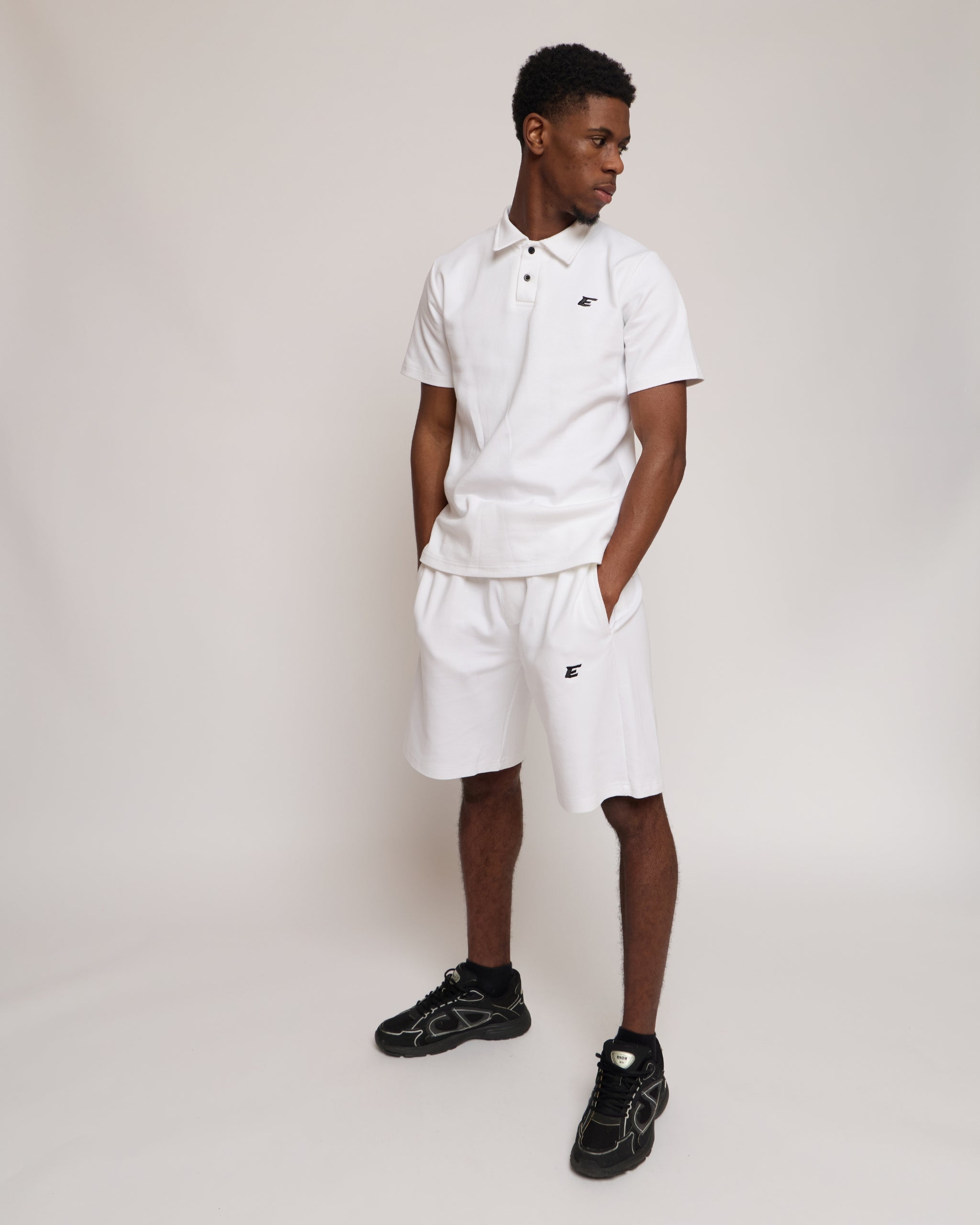 Man wearing a white polo shirt and shorts with an 'E' logo, standing against a plain background