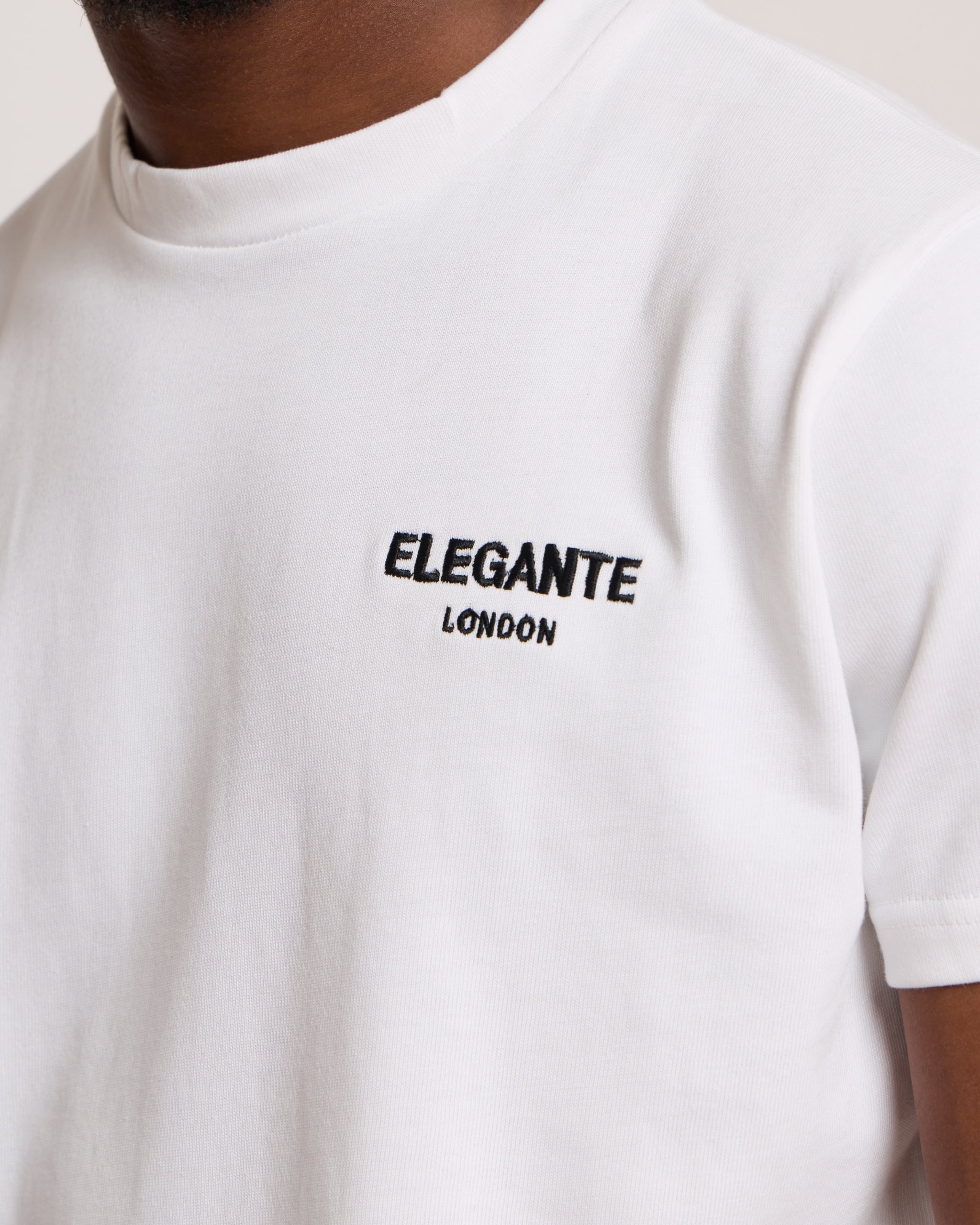 Man wearing a white cotton t-shirt with 'Elegante London' logo on a plain background