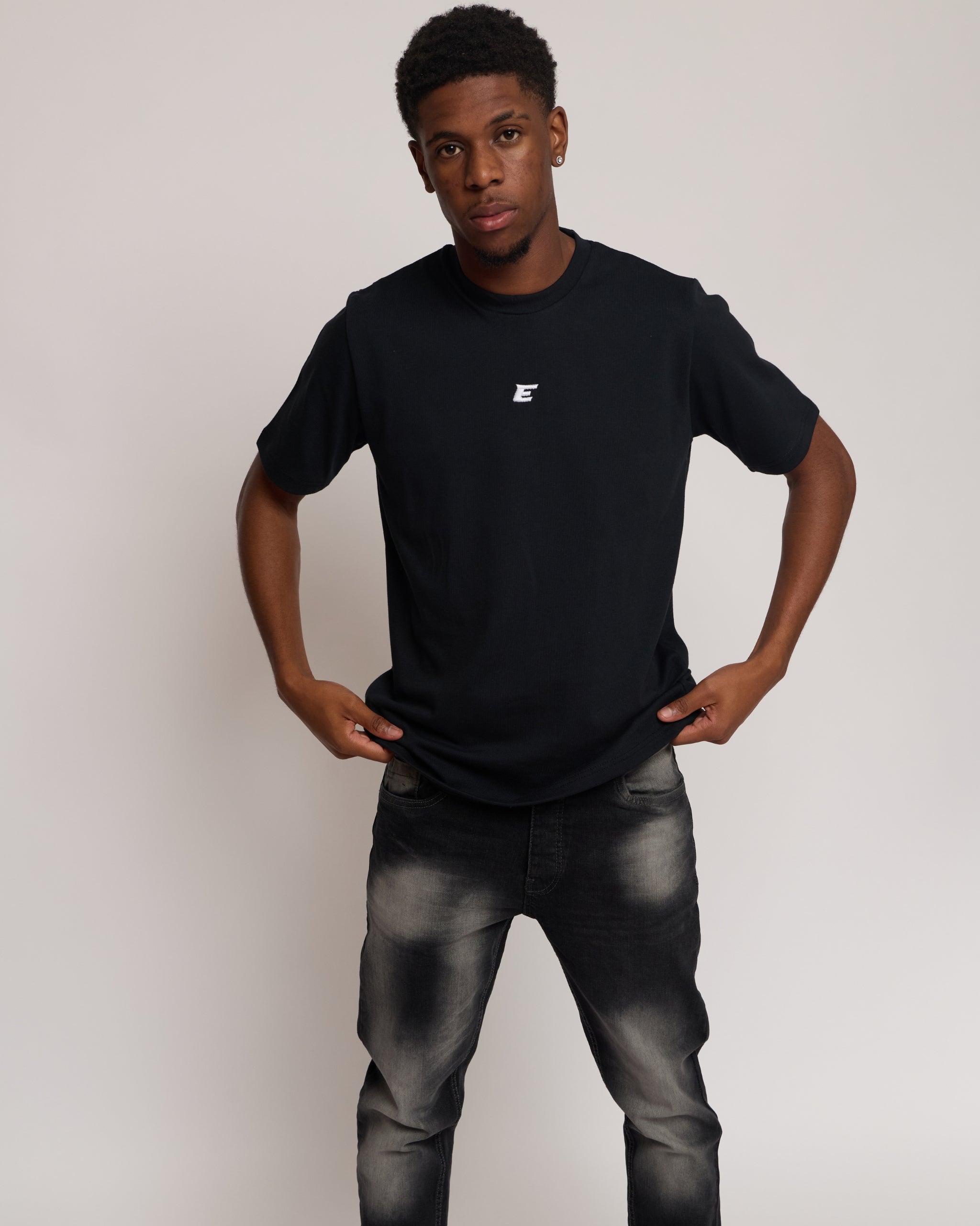Man wearing a black t-shirt with a small white 'E' logo and dark jeans on a light gray background