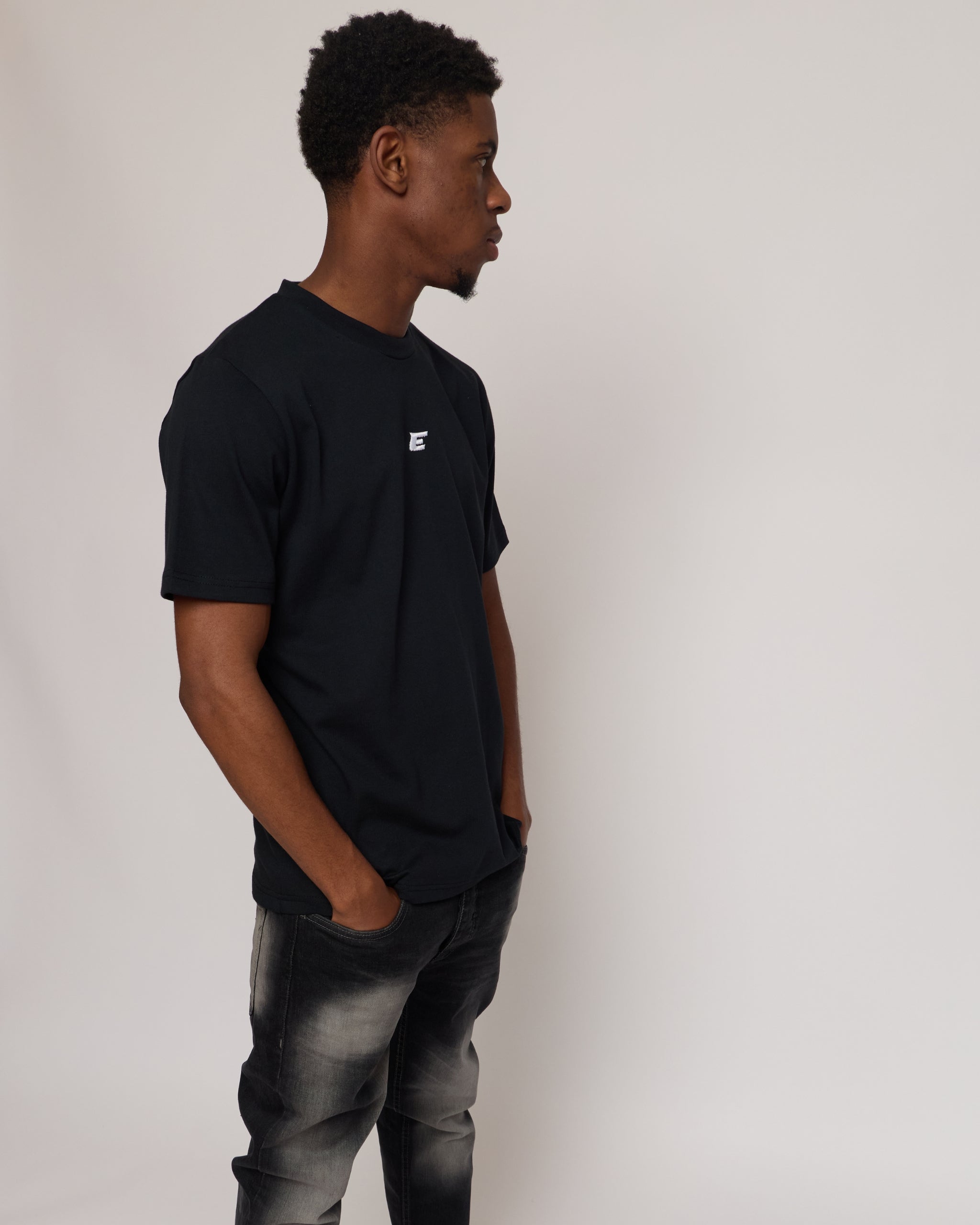 man wearing a black cotton t-shirt with 'E' Logo and grey jeans on a white background
