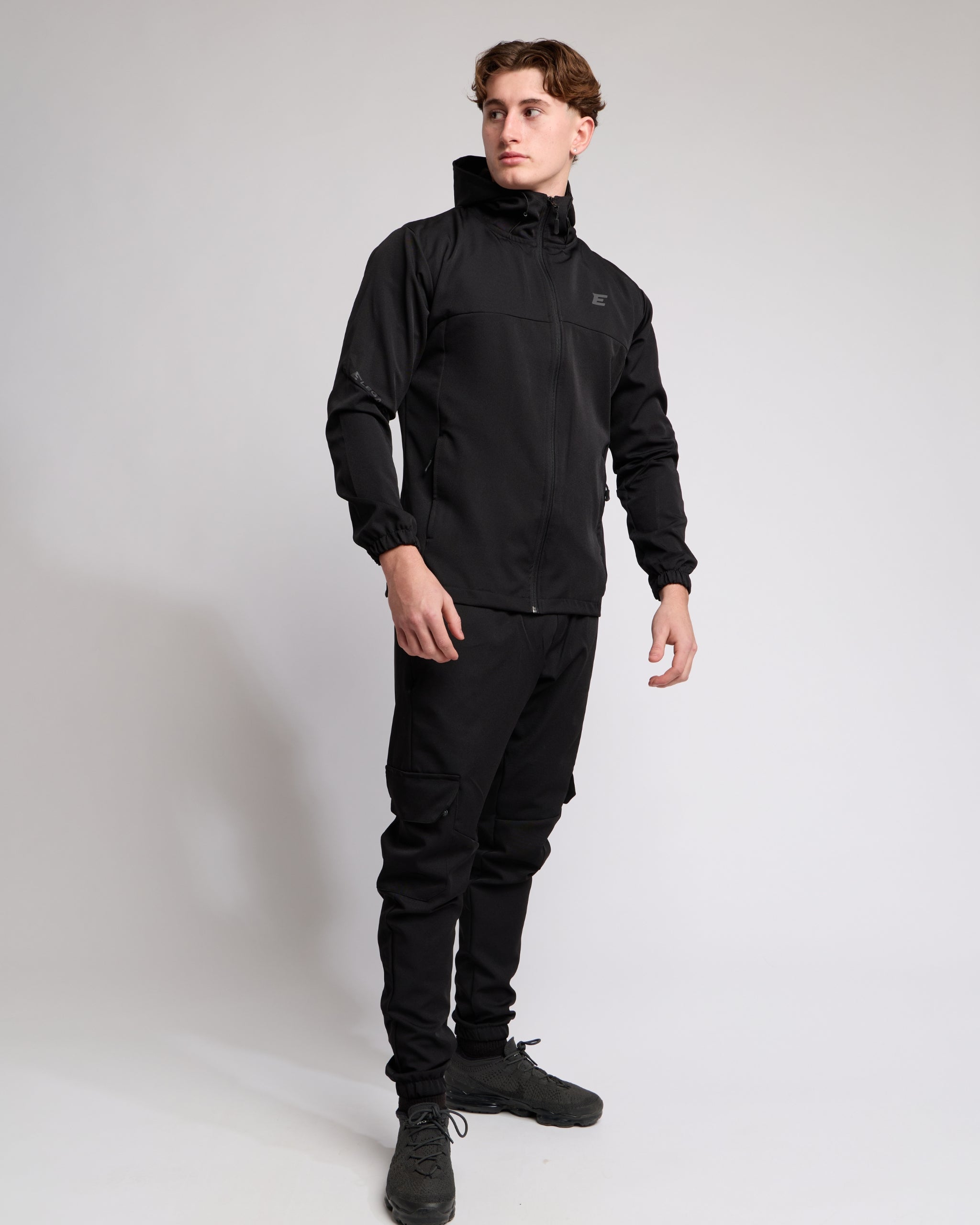 Man wearing an all Black sports tracksuit jacket with hood and trousers on a white background