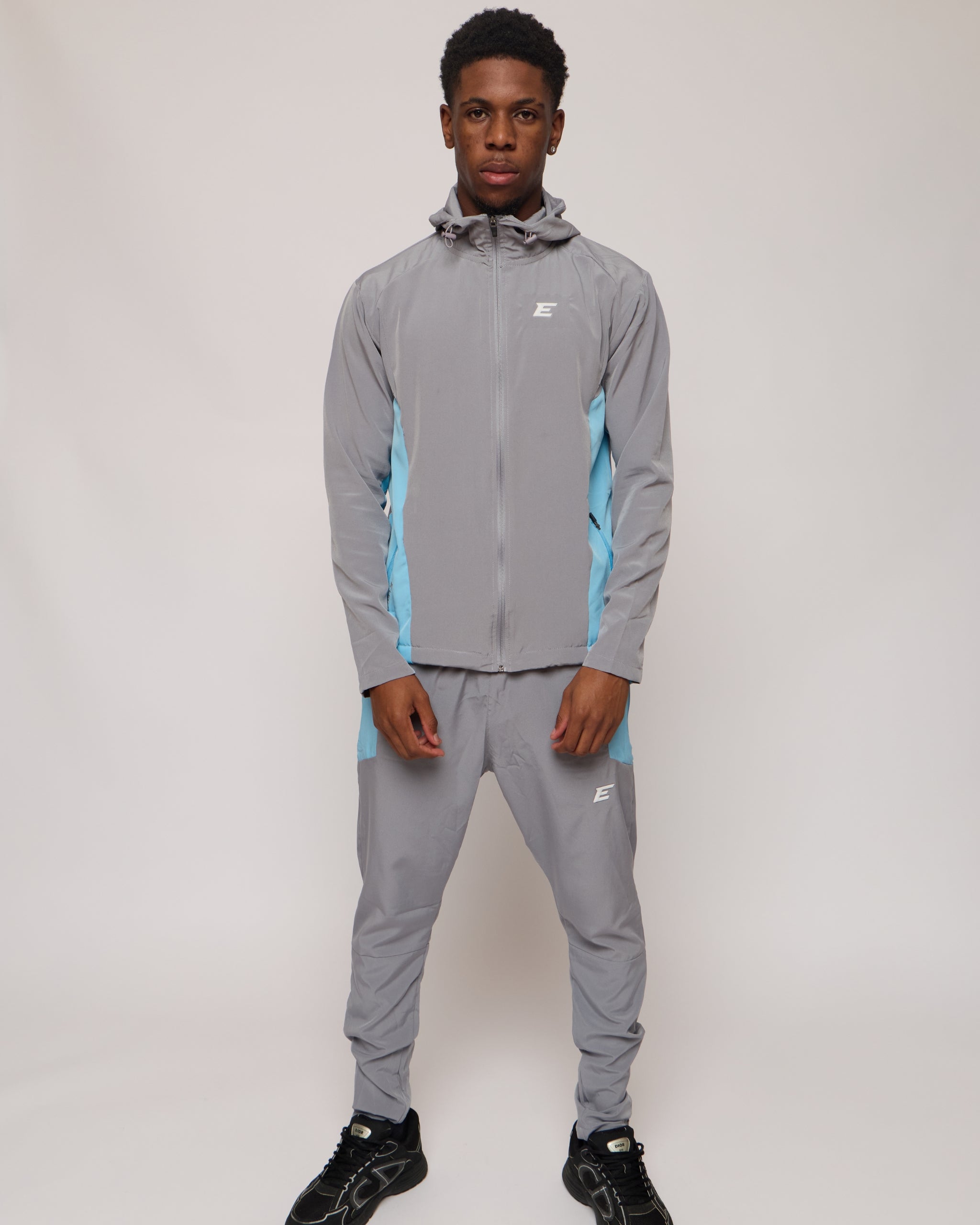 Man wearing a grey and blue tracksuit jacket with hood and matching joggers with an E logo on a plain background