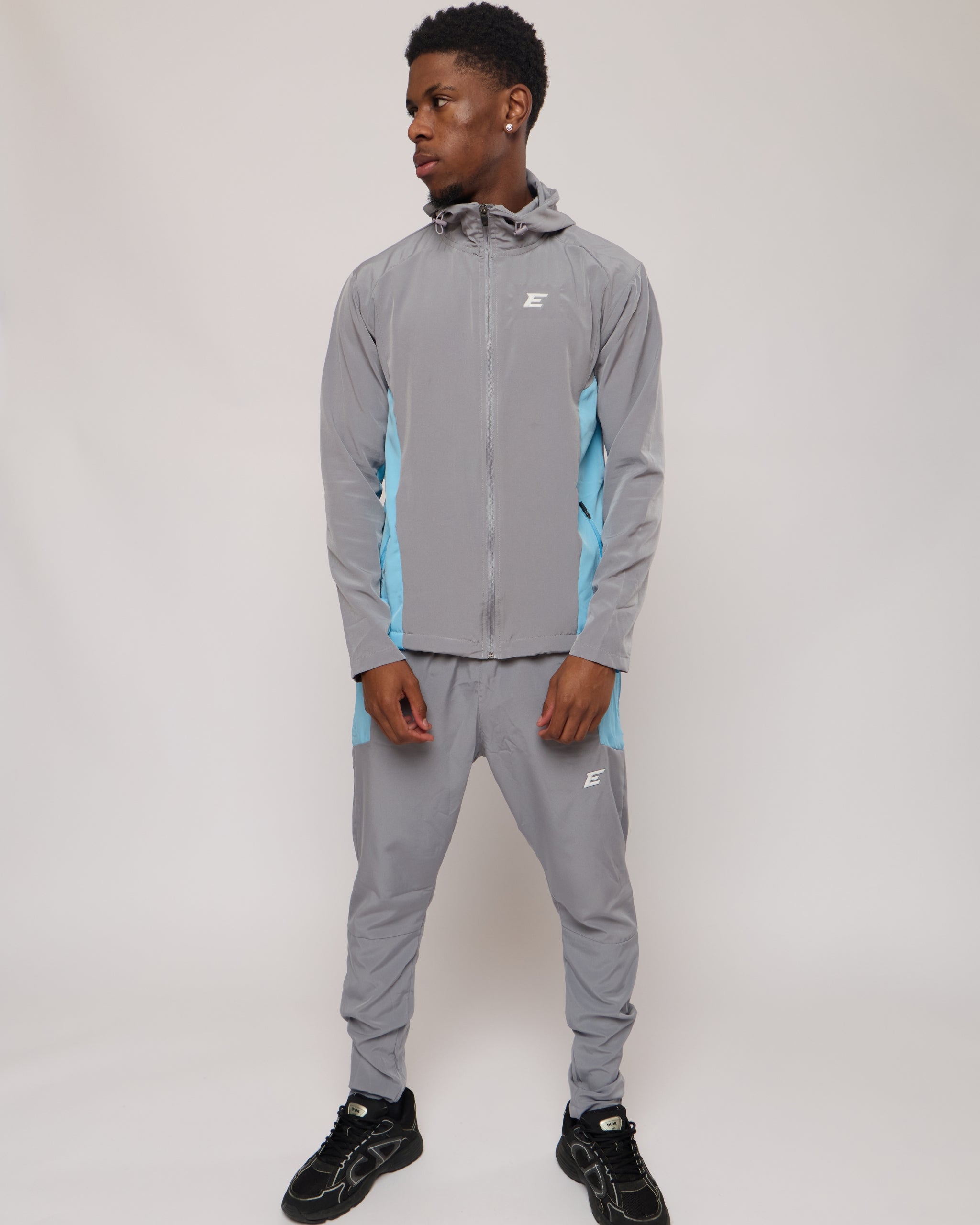 Man wearing a grey and blue tracksuit jacket with hood and matching joggers with an E logo on a plain background