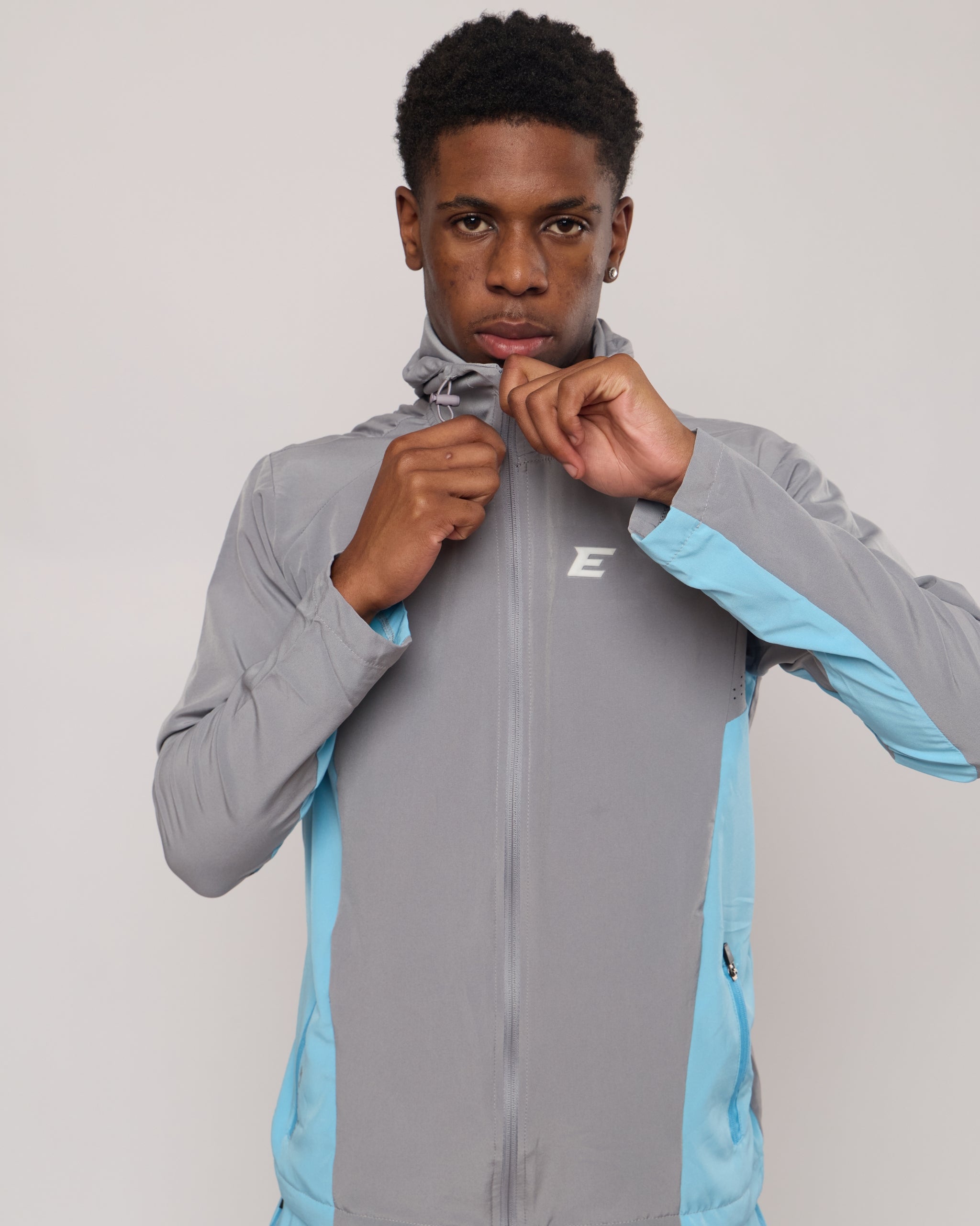 Man wearing a grey and blue tracksuit jacket with hood with an E logo on a plain background