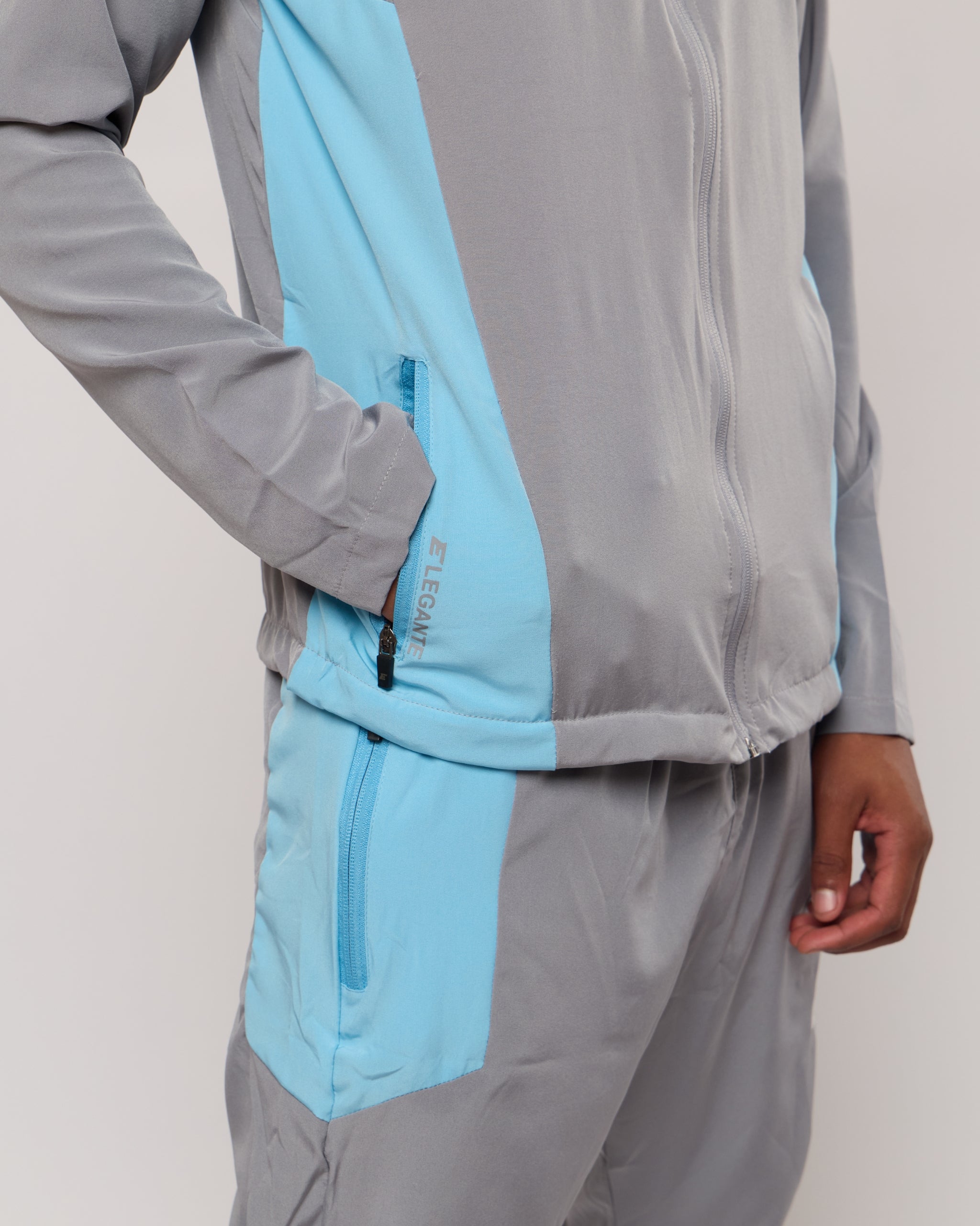 Man wearing a grey and blue tracksuit jacket with zip pockets with an Elegante logo on a plain background