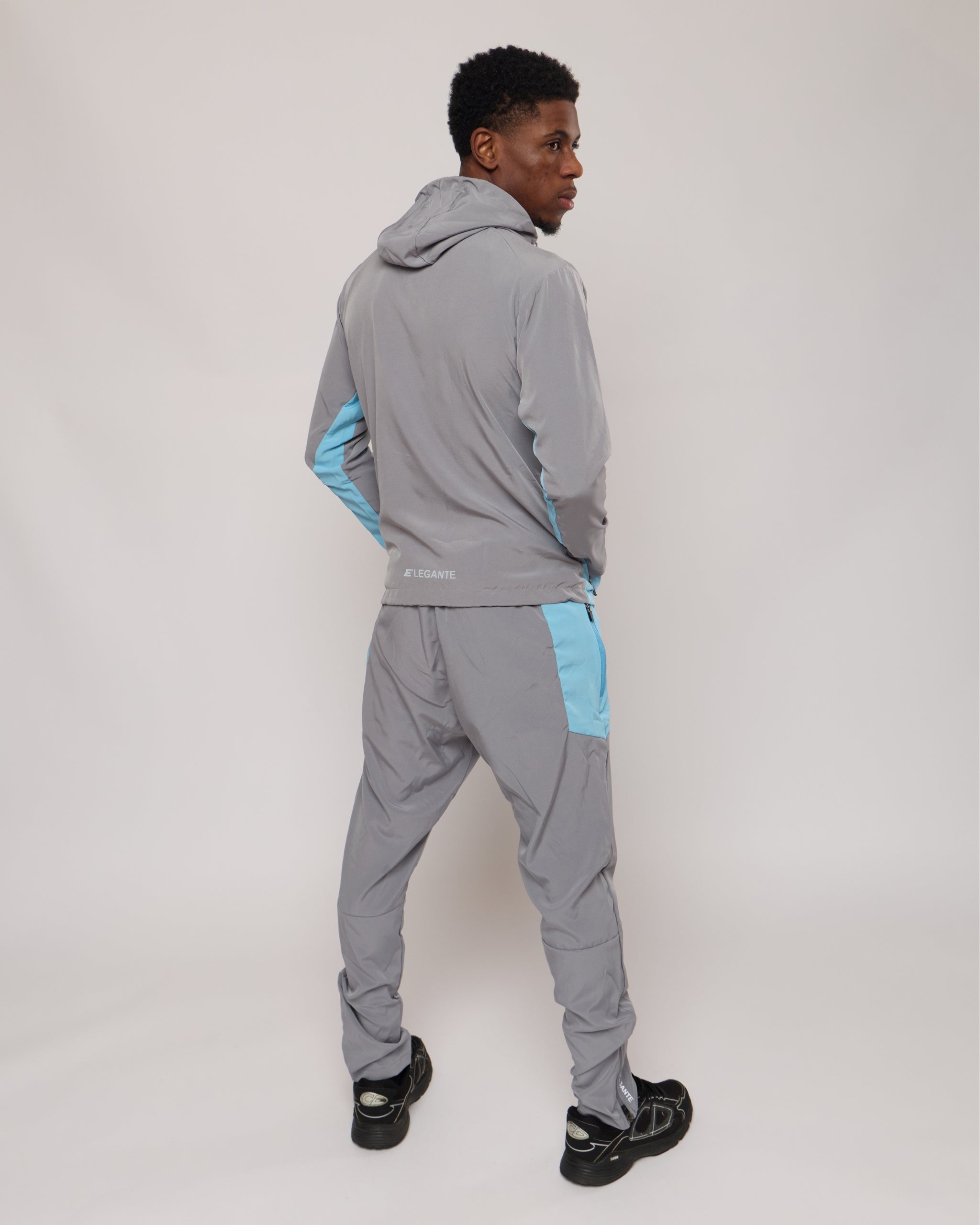 Man wearing a grey and blue tracksuit jacket with hood and matching joggers with an E logo on a plain background