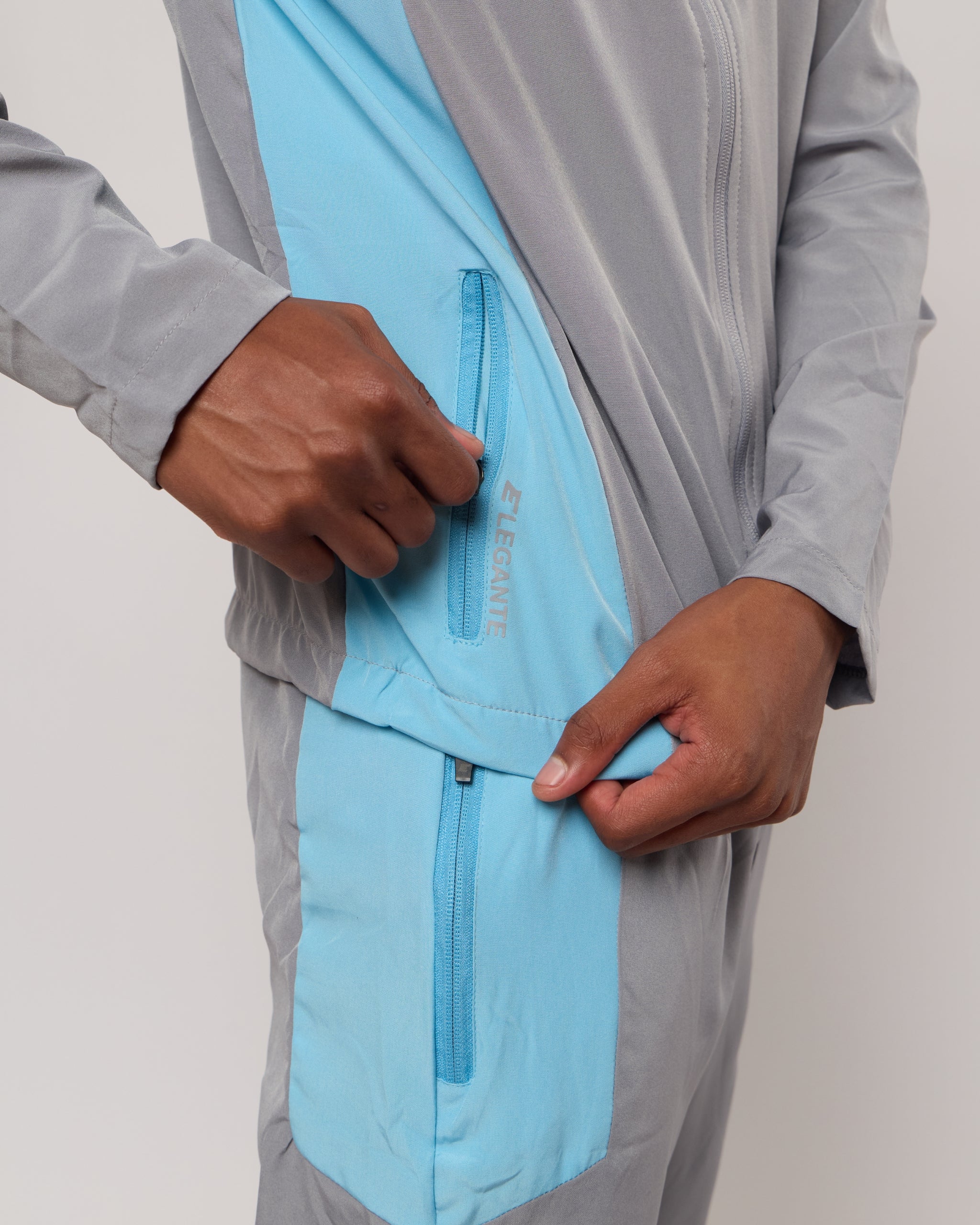 Man wearing a grey and blue tracksuit jacket with zip pockets with an Elegante logo on a plain background