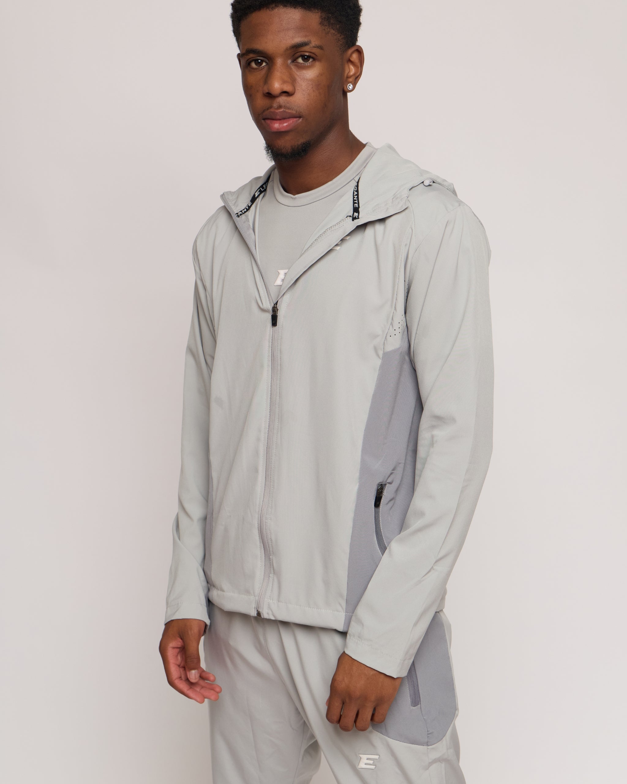 Man wearing a light gray jacket with a hood and matching joggers on a plain background