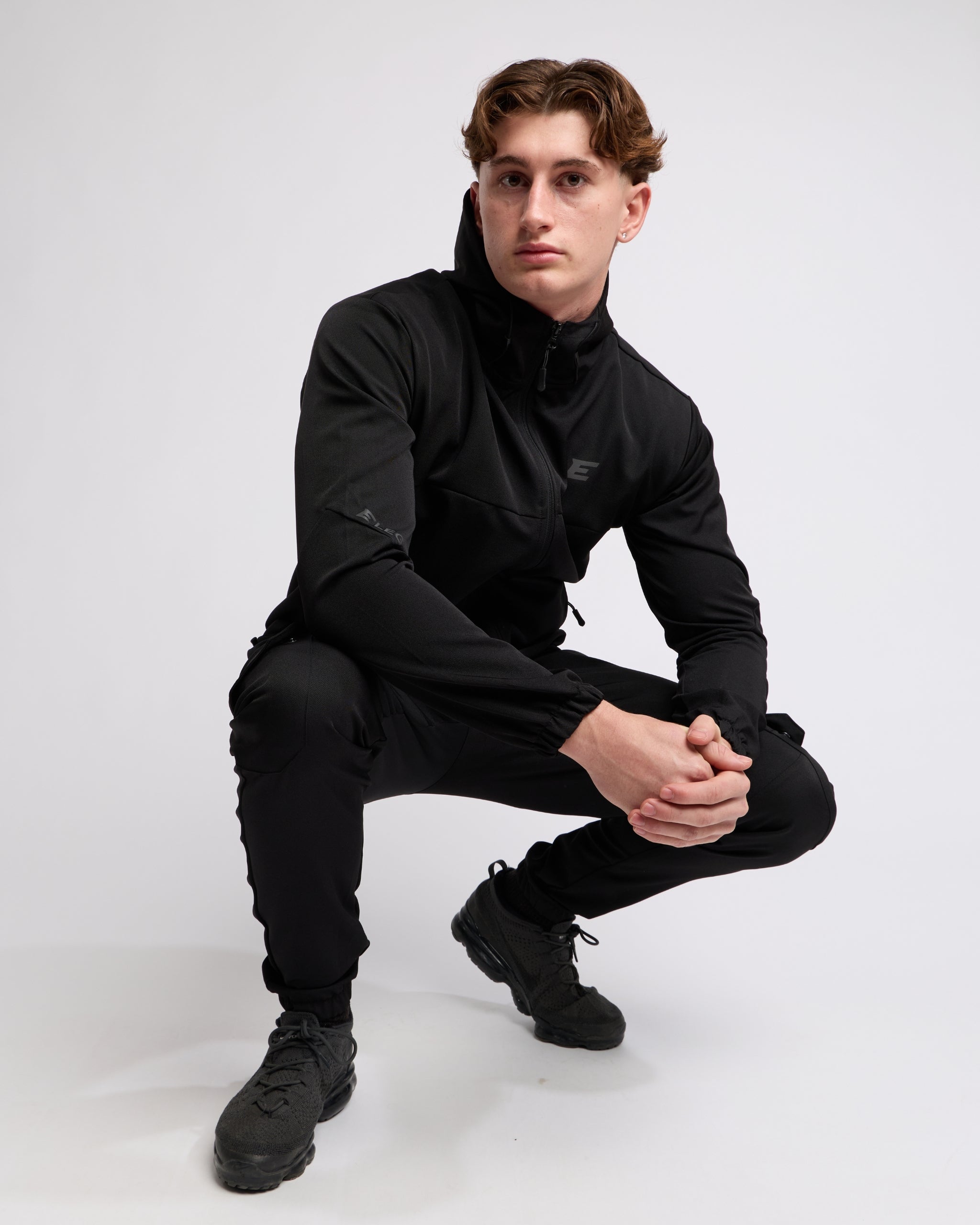 Man wearing an all Black sports tracksuit jacket with hood and trousers on a white background