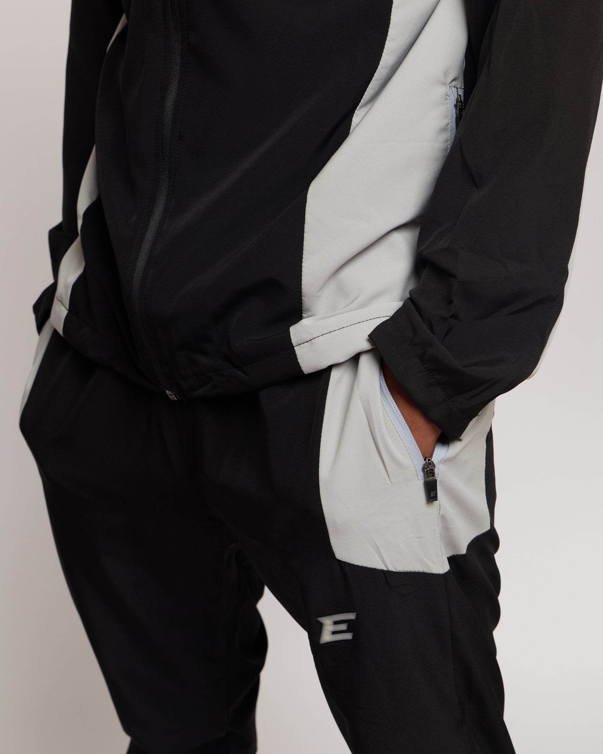 Man wearing a black and white tracksuit jacket with hood with matching joggers and an E logo on a plain background