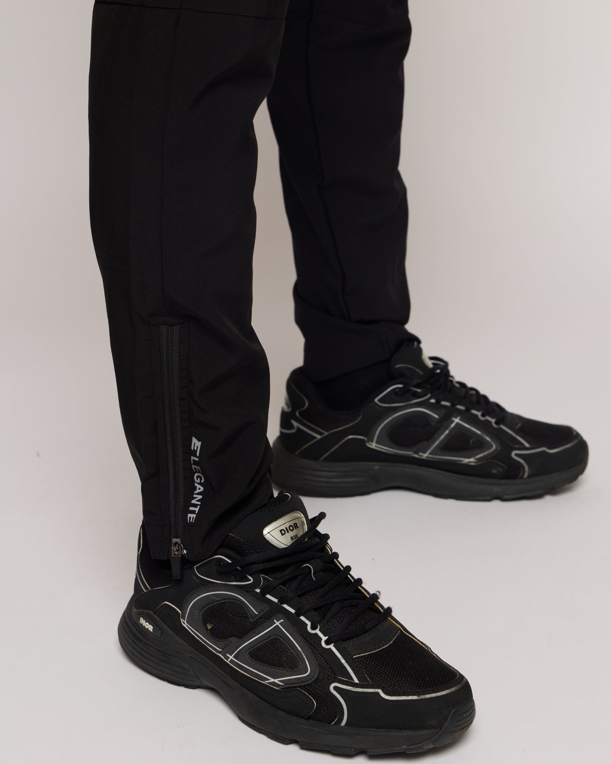man wearing black tracksuit bottoms with elegante brand logo with black trainers on a white background