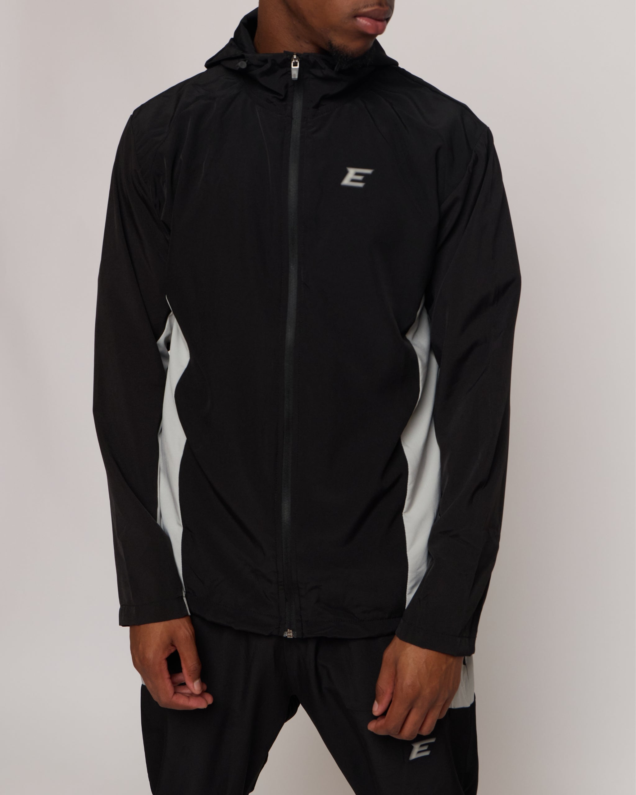 Man wearing a black and white tracksuit jacket with hood with matching joggers and an E logo on a plain background