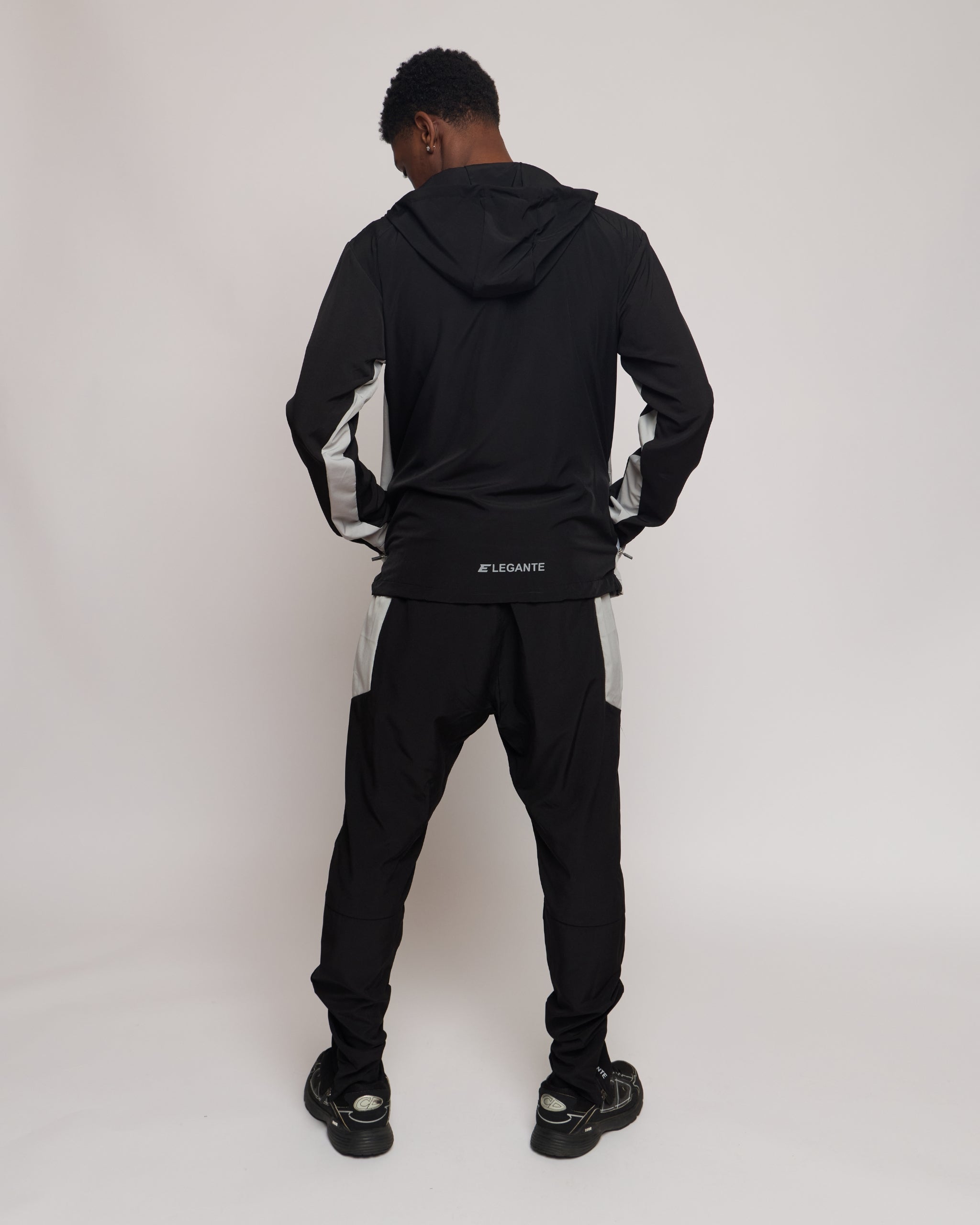 Man wearing a black and white tracksuit jacket with hood with matching joggers and an elegante logo on a plain background