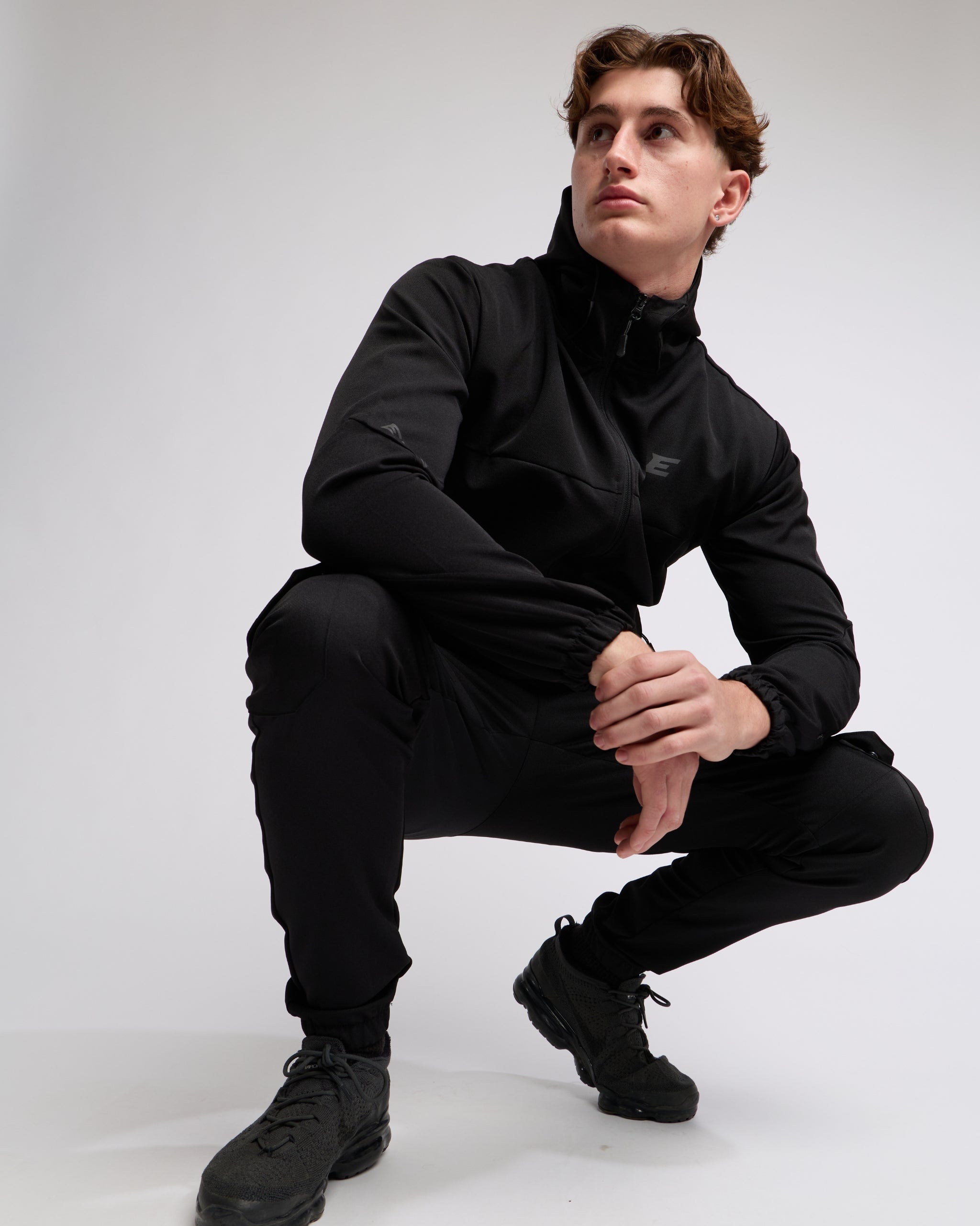 Man wearing an all Black sports tracksuit jacket with hood and trousers on a white background
