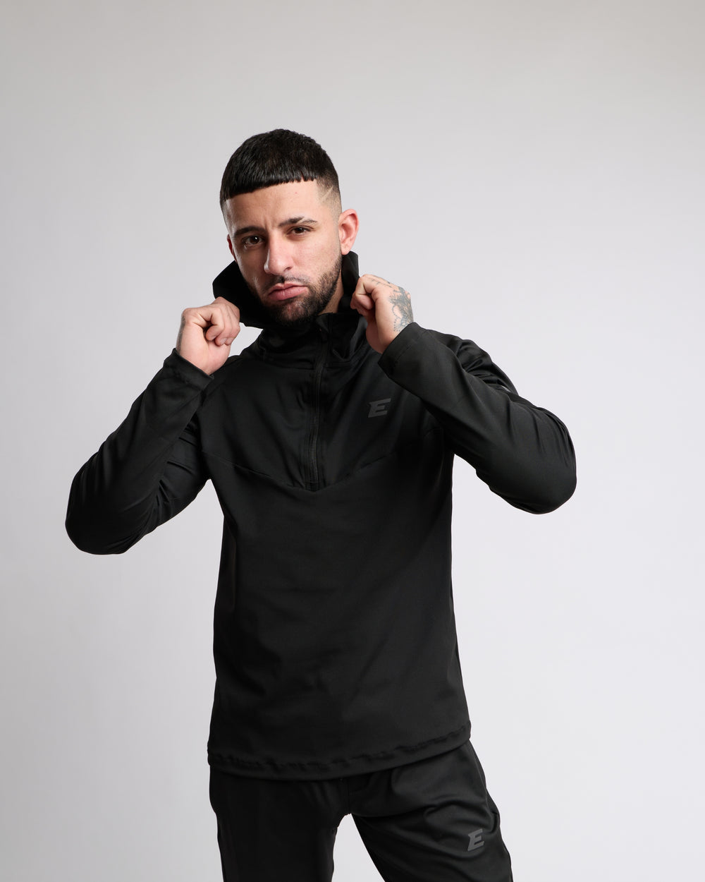 Man wearing an all black tracksuit zip top with hood against a gray background