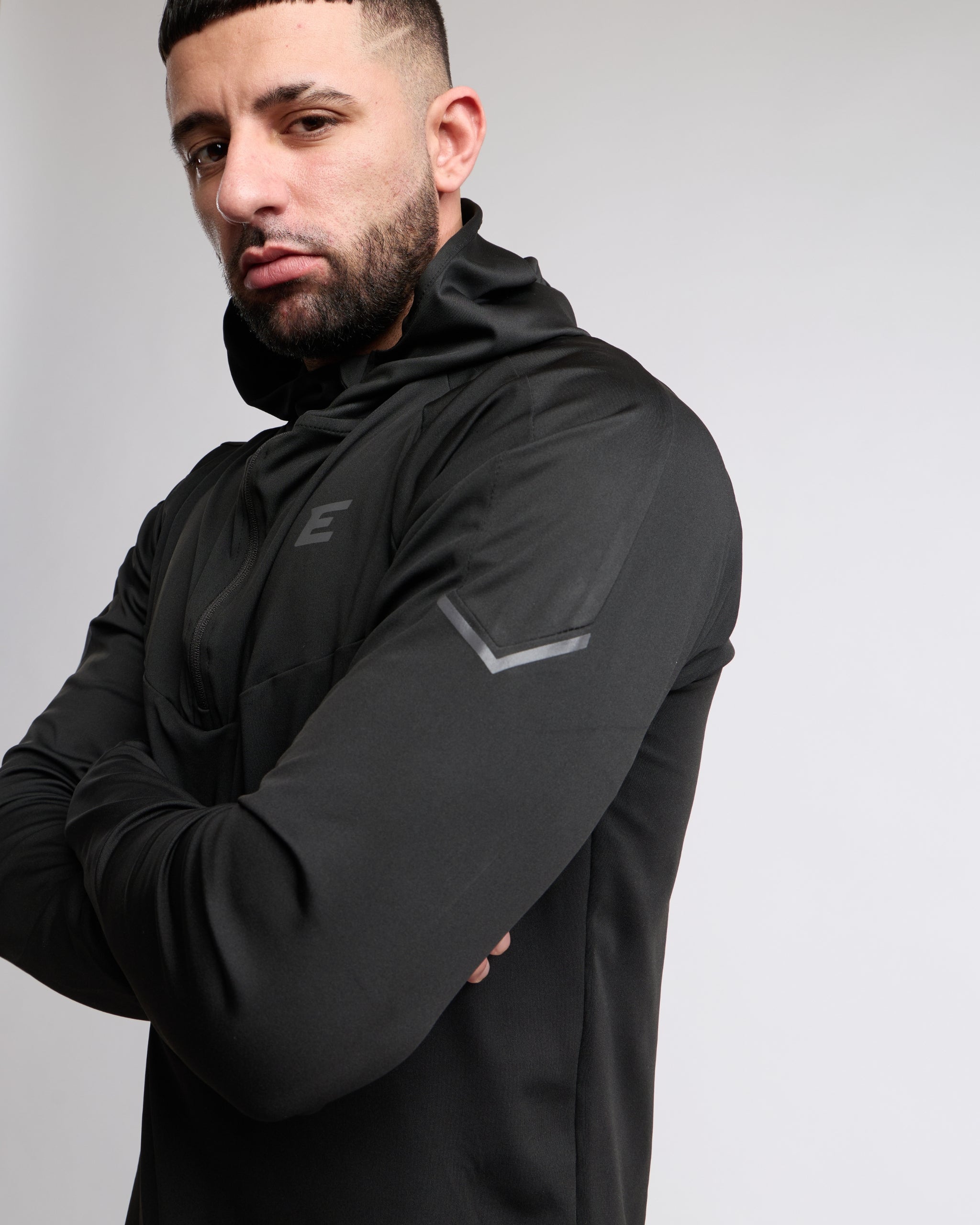 Man wearing an all black tracksuit zip top with hood against a gray background
