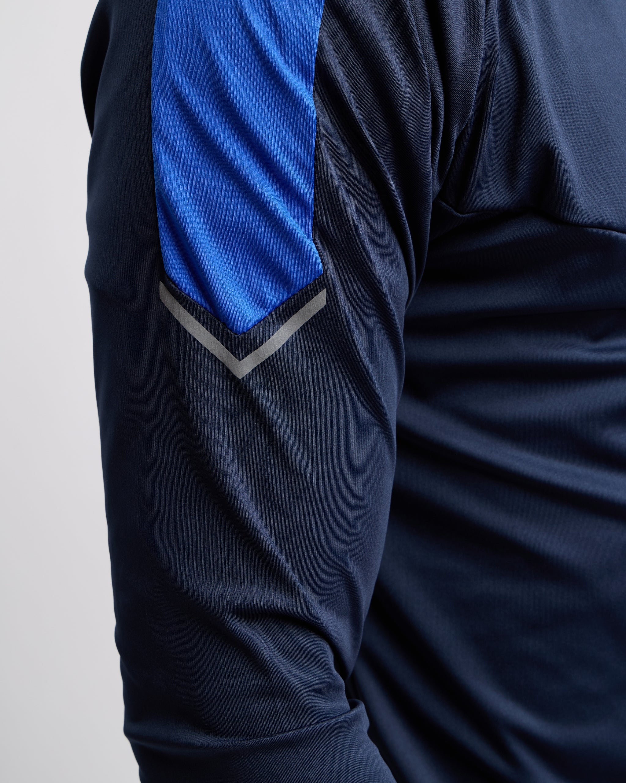 Man wearing a navy and blue athletic zip top with reflective ascent on the sleeve on a gray background