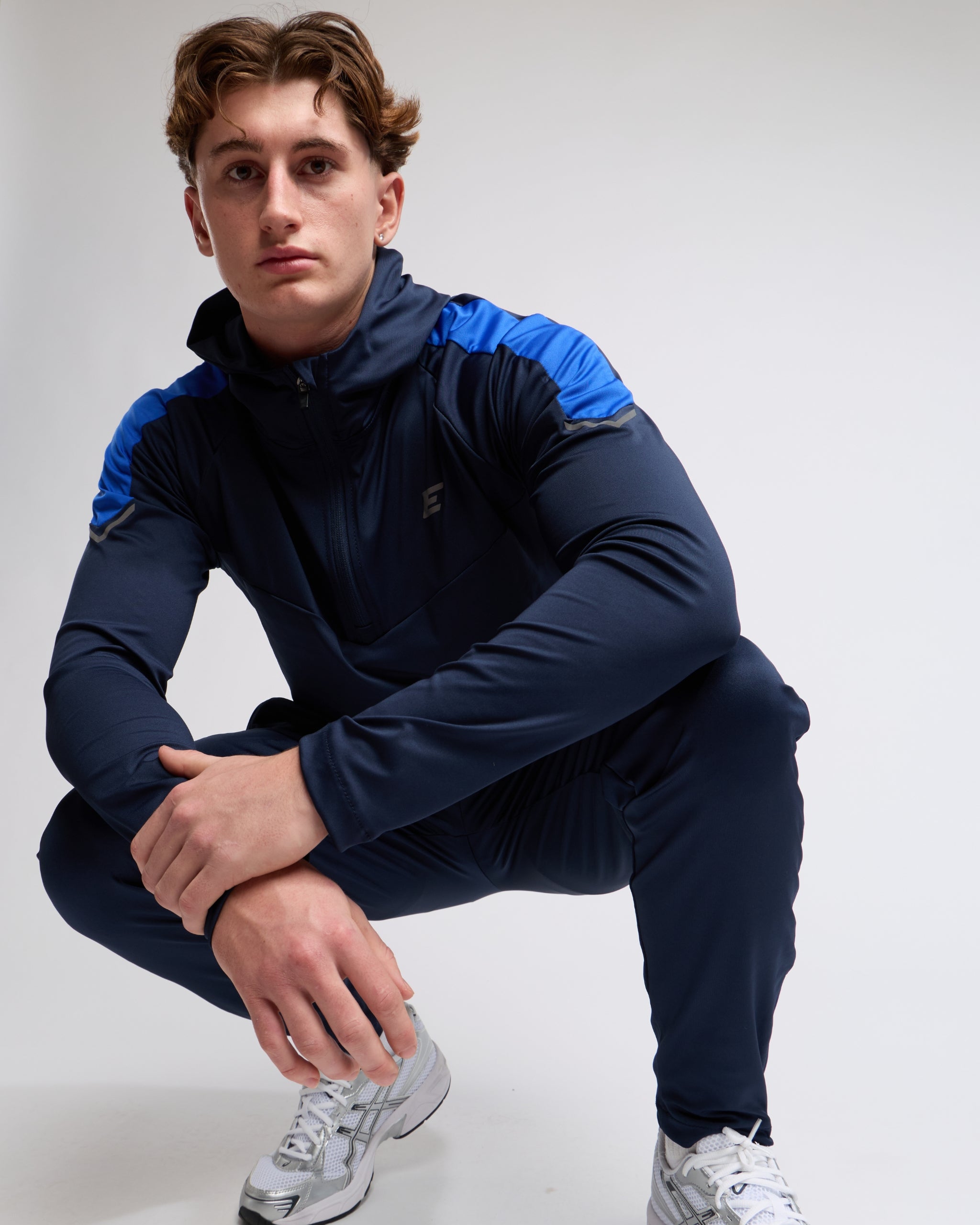 Man wearing a navy and blue athletic zip top with joggers on a gray background