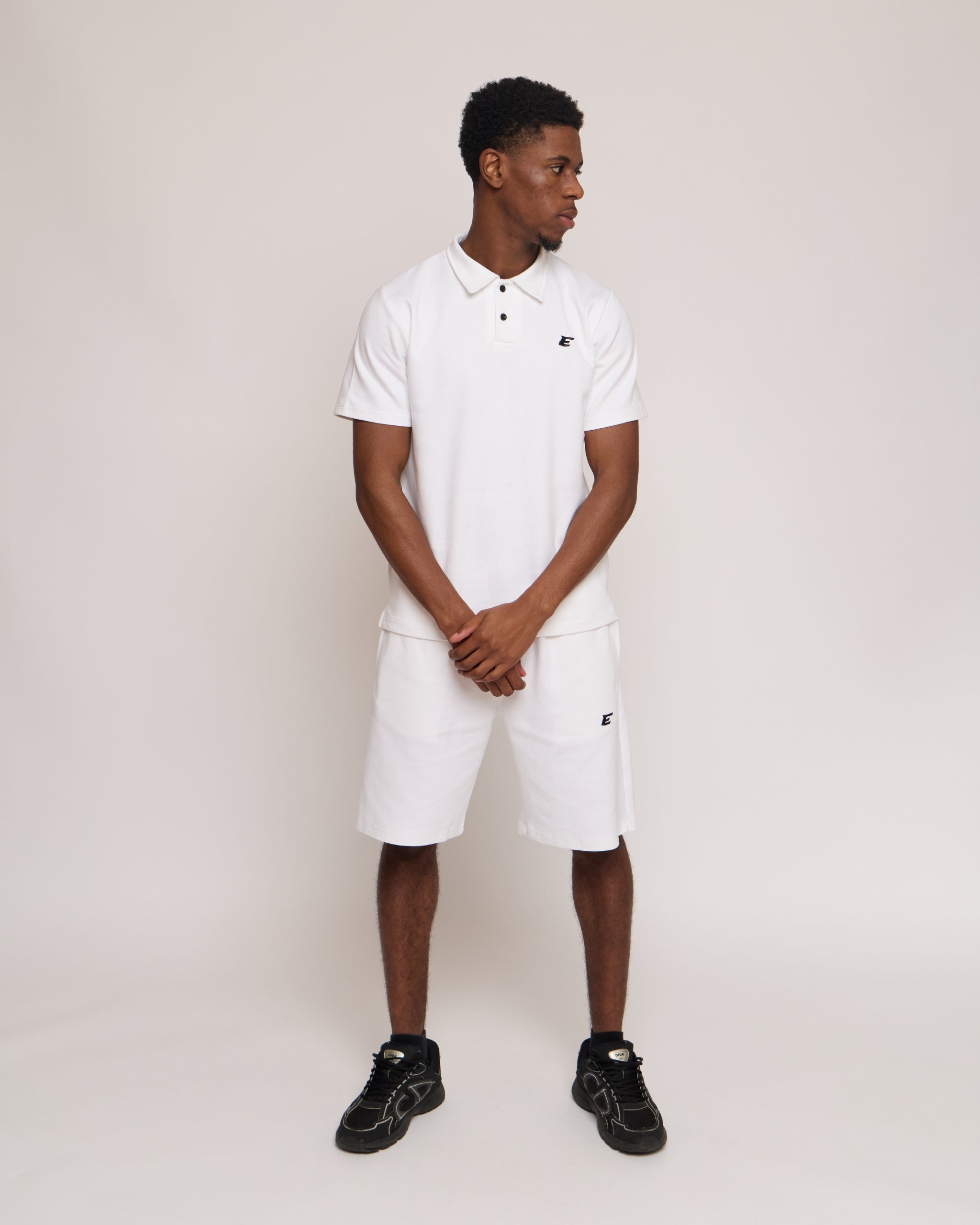 Man wearing a white polo shirt and shorts with an 'E' logo, standing against a plain background