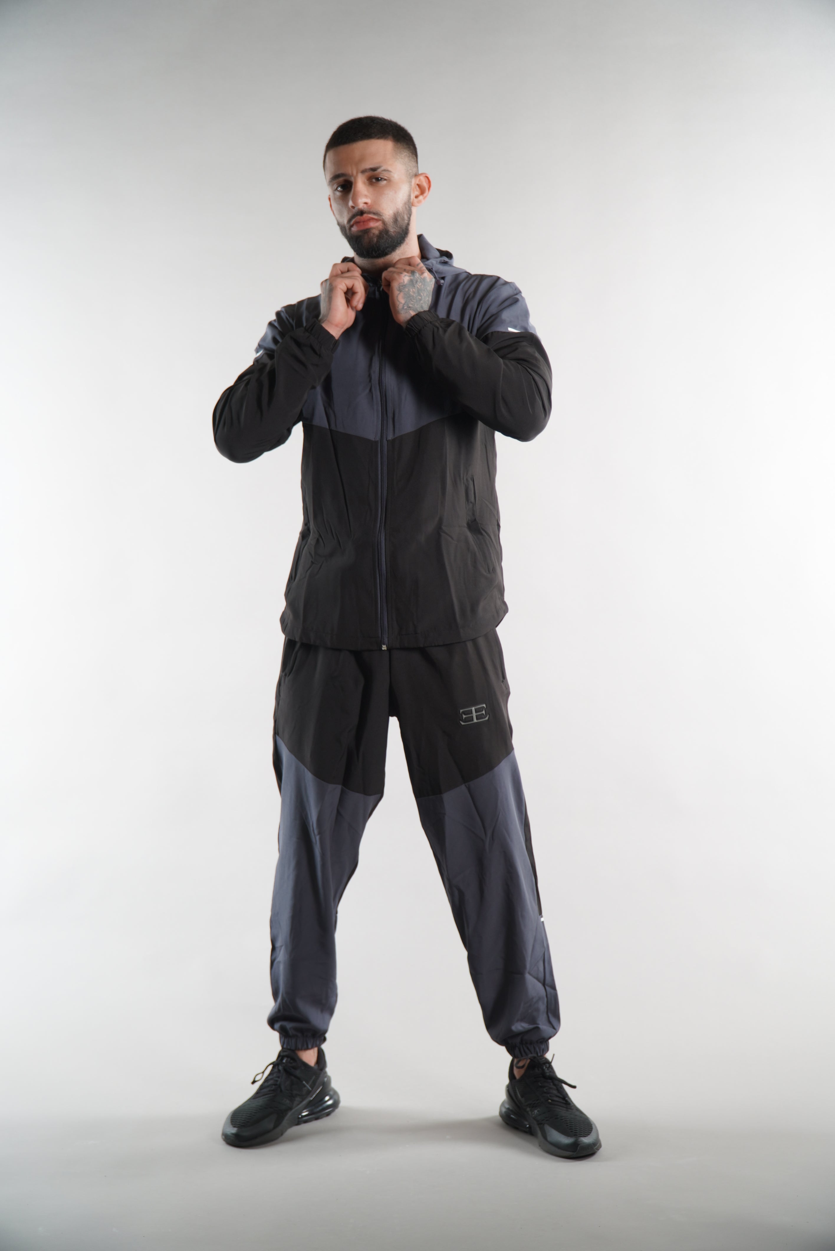 Man wearing black and dark grey tracksuit Jacket with hood and tracksuit pants set, with his hands on the hood, on white background, sports clothing for men