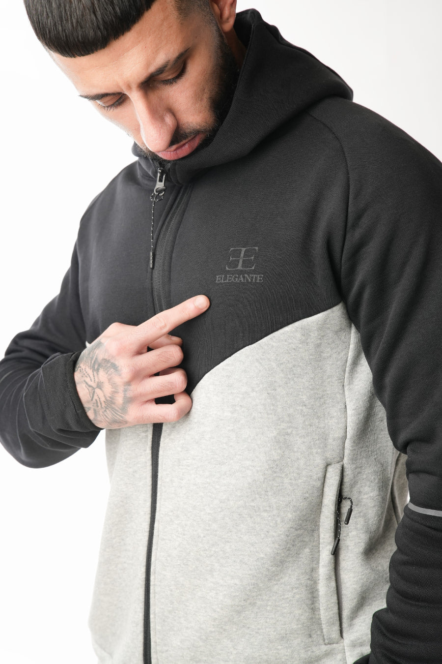 Man wearing a black and gray hoodie with elegante brand logo, pointing to it.