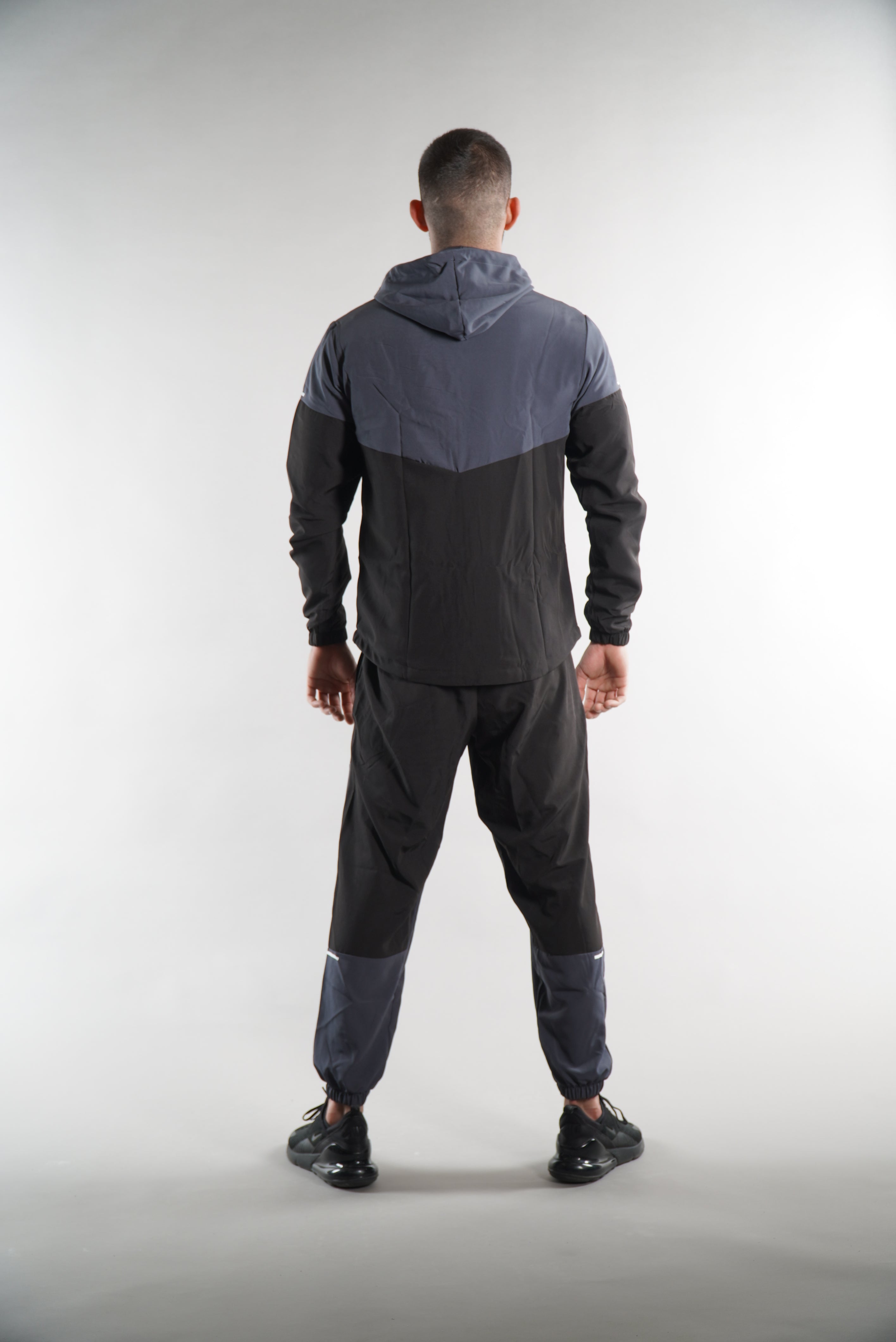 Man wearing black and dark grey tracksuit Jacket with hood and tracksuit pants set, view form the back, on white background, sports clothing for men