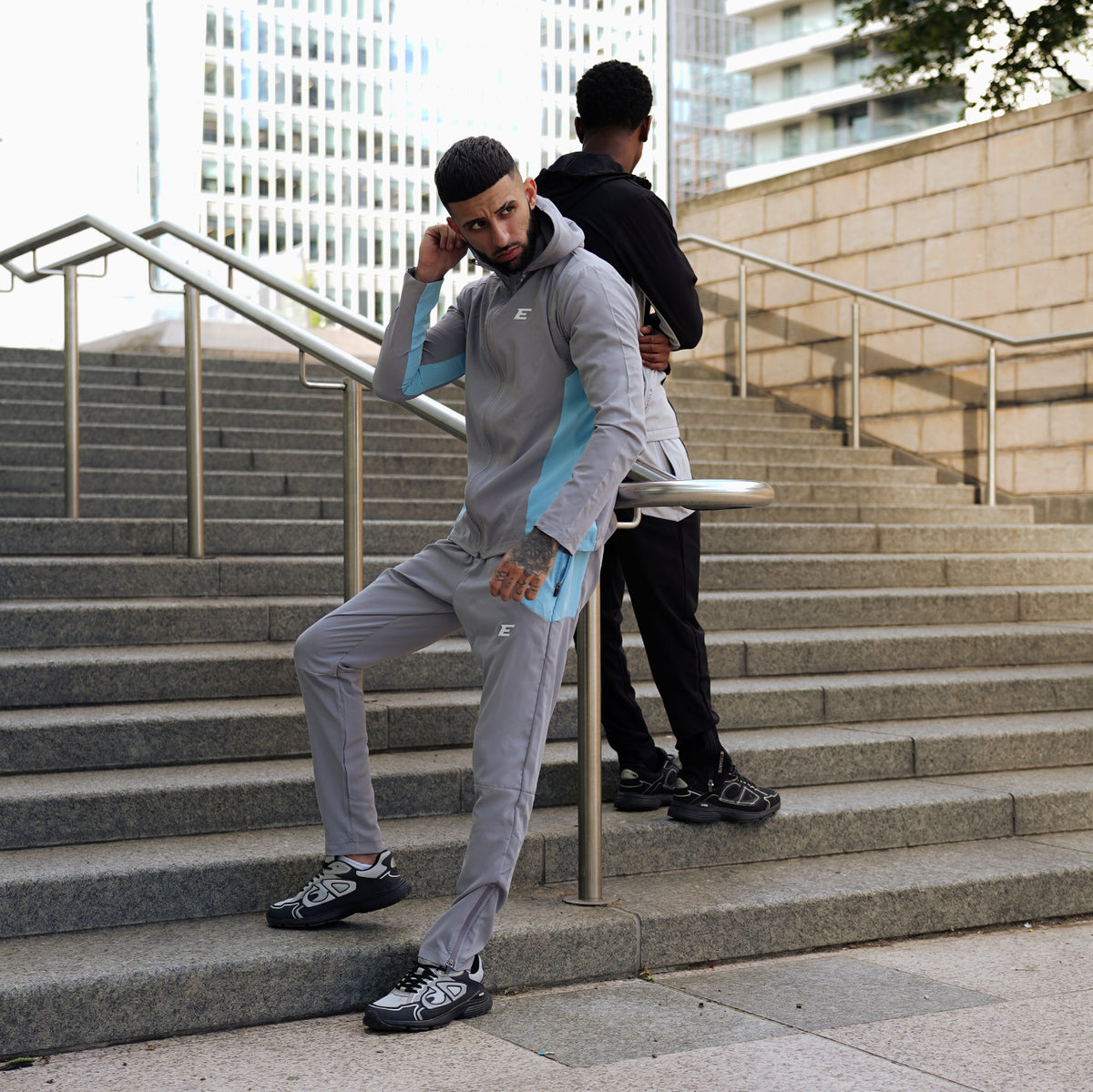 two men wearing sports tracksuits on city stairs one man wearing a grey and light blue tracksuit set and another man wearing a black and grey tracksuit set