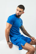 Man wearing a blue matching athletic shorts and tshirt sitting on a white surface with a plain background
