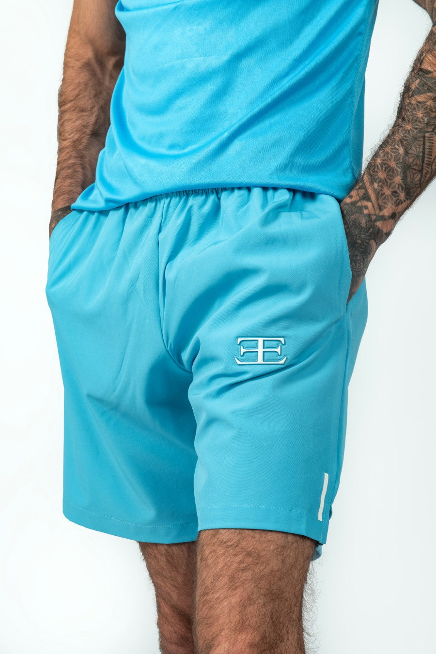 man wearing a matching blue athletic shorts and tshirt outfit with elegante logo with hands in pockets on a white background