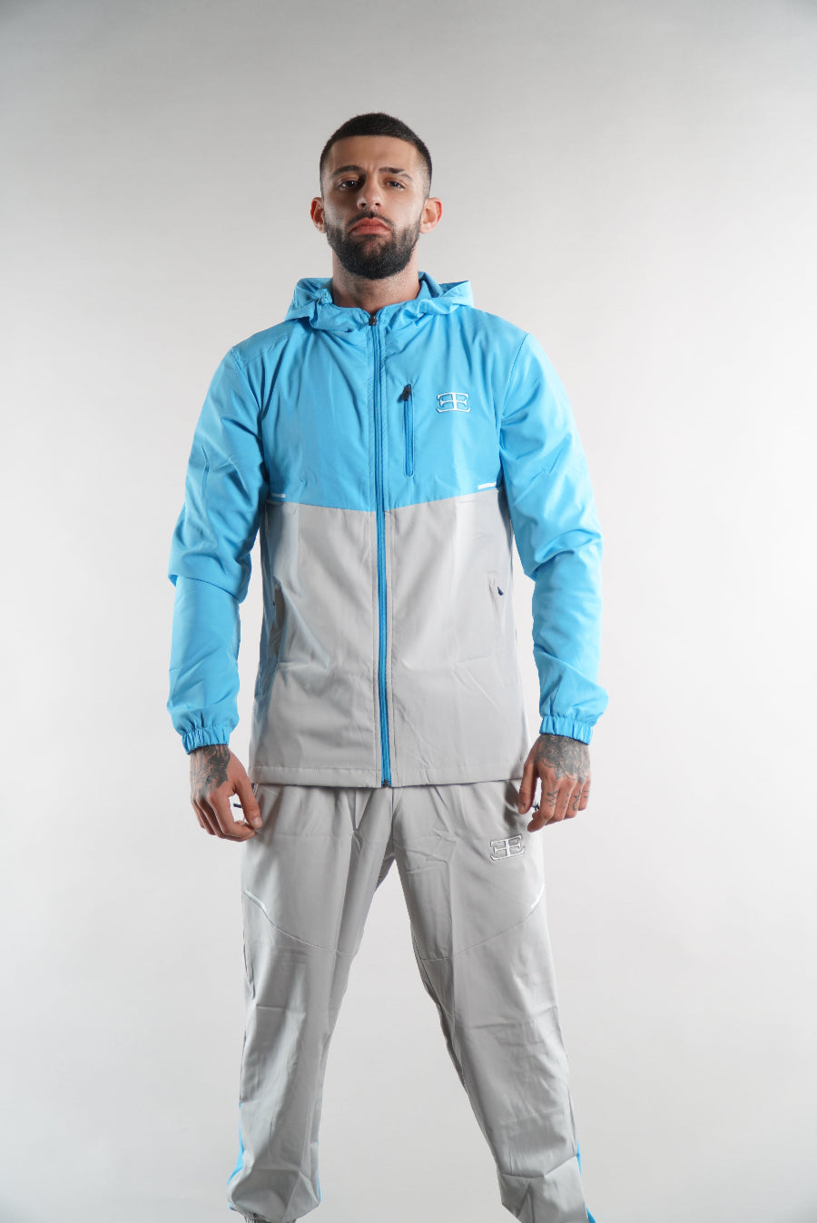 man wearing Elegante branded turquoise blue Tracksuit jacket with hood and grey blue joggers on white background