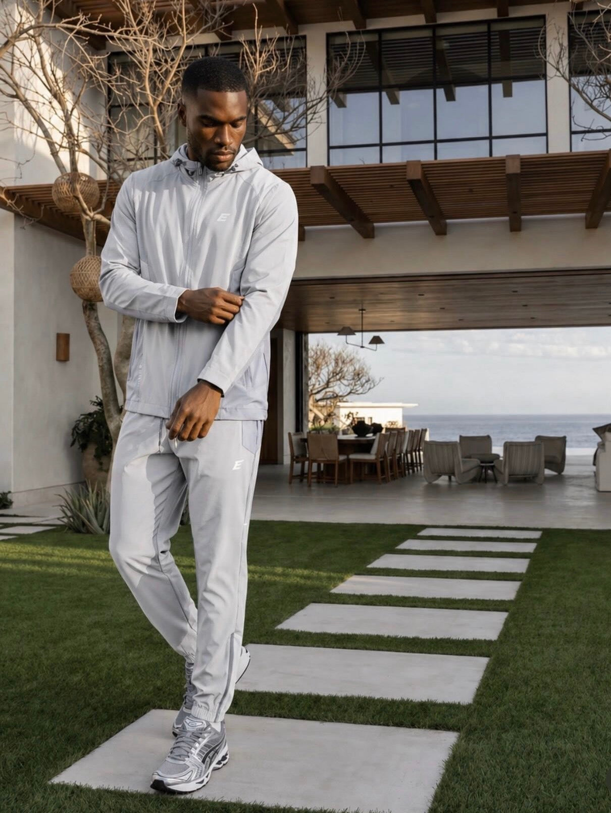 Man in a light gray tracksuit set standing in a modern outdoor setting with ocean view