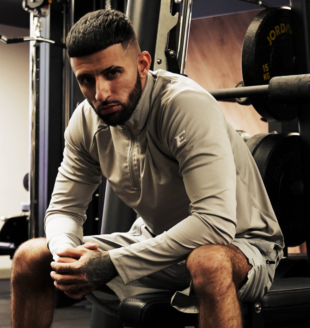 A man in a gym setting wearing a light gray long-sleeve athletic quarter-zip pullover and matching shorts, sitting on a weight bench in front of gym equipment.