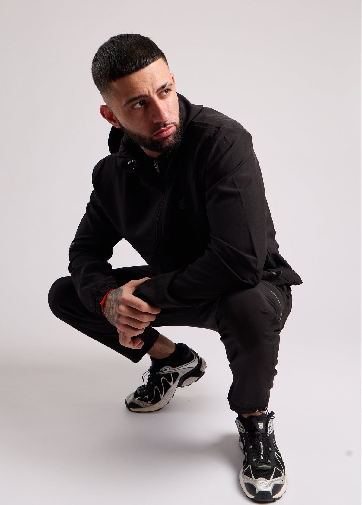 A man crouching in a studio setting, wearing a black zip-up hoodie, black pants, and black and white sneakers against a light gray background.