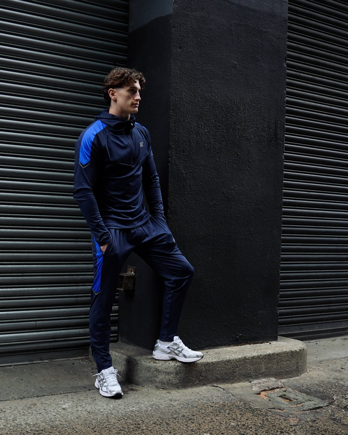 man wearing a sporty blue tracksuit leaning against a dark wall on a street.