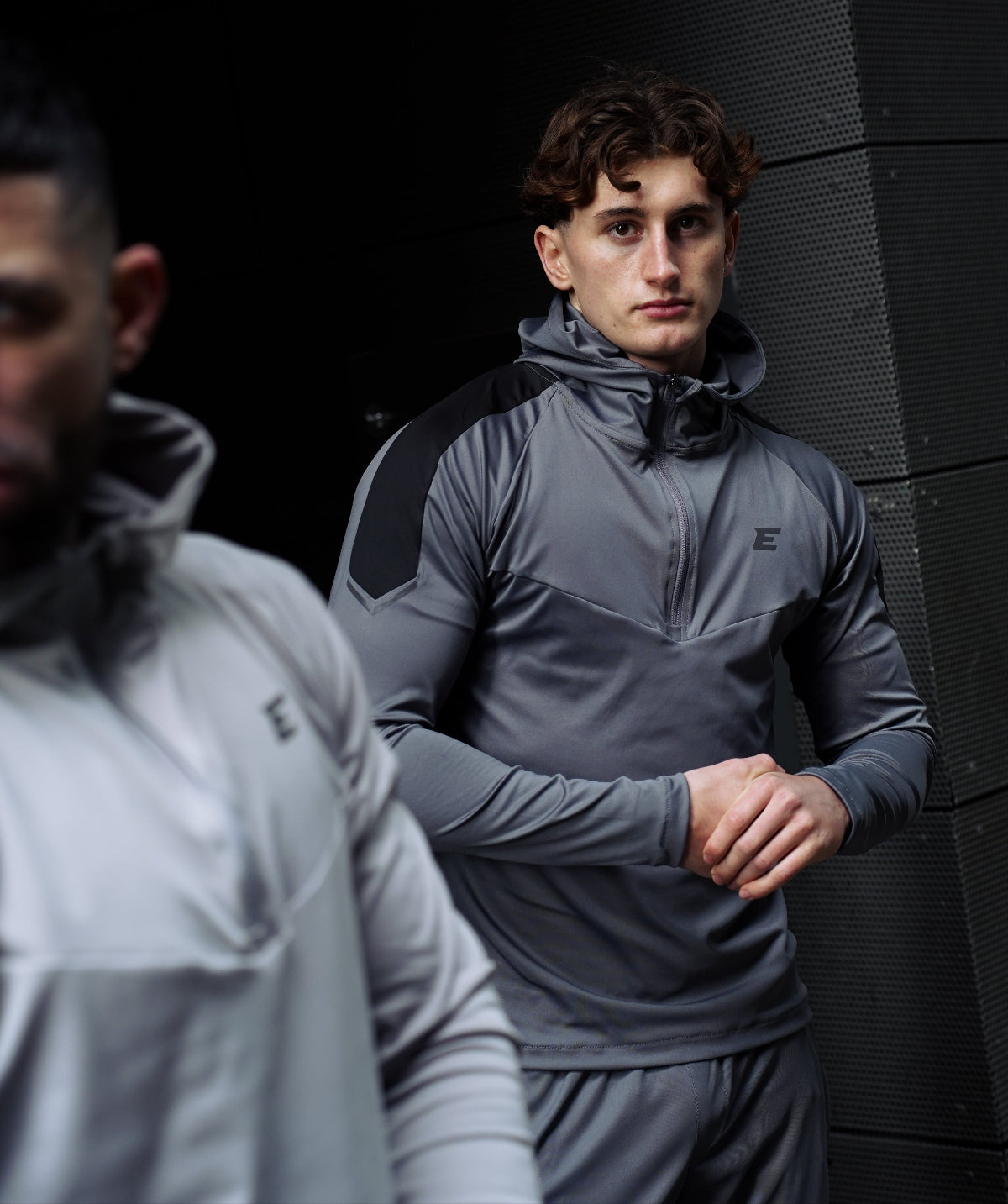 Two men wearing gray athletic jackets standing against a dark wall.