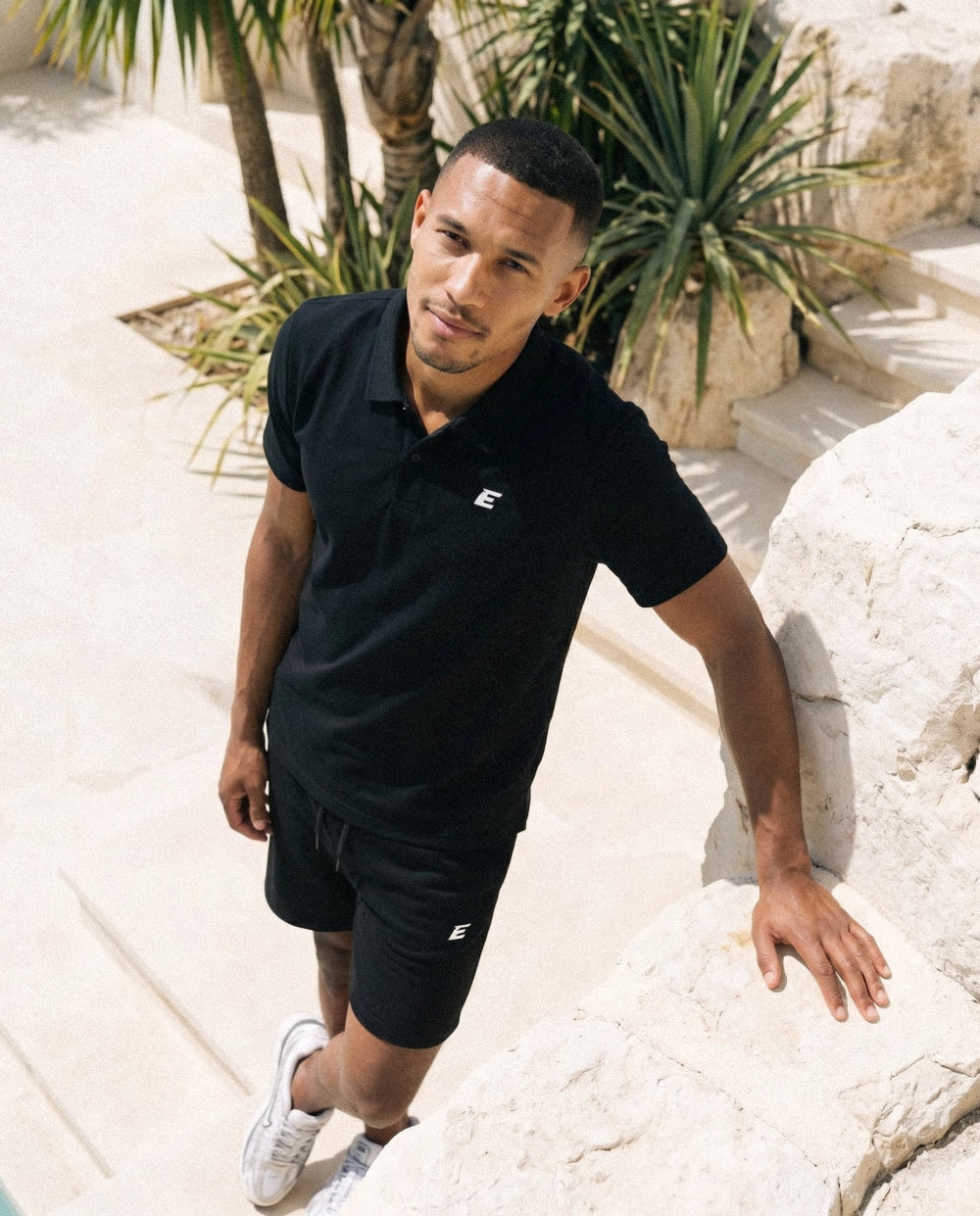 Man wearing a black polo tshirt and shorts with a E logo, standing sunny background.