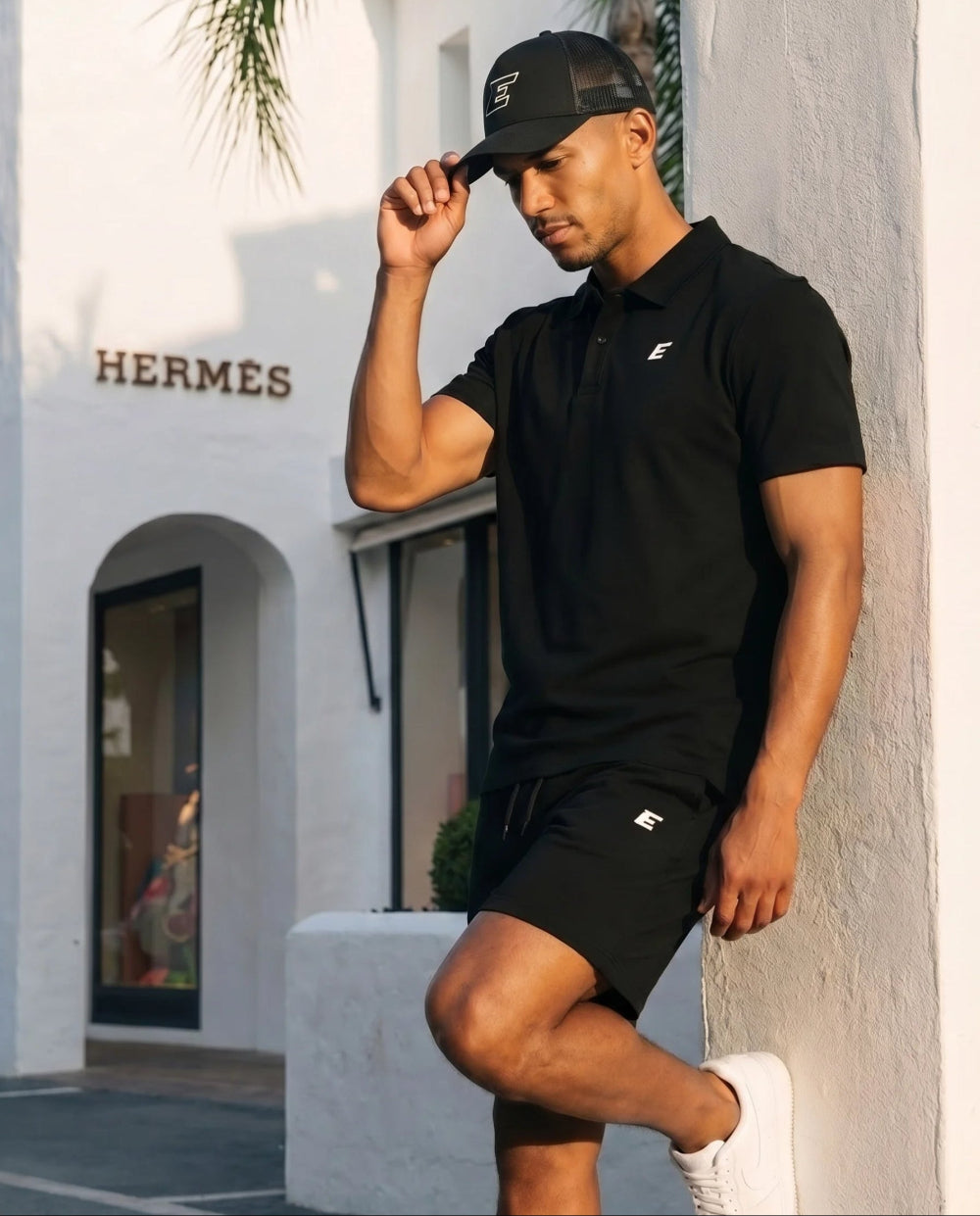 Man wearing a black polo shirt and shorts with a cap, standing in front of a building
