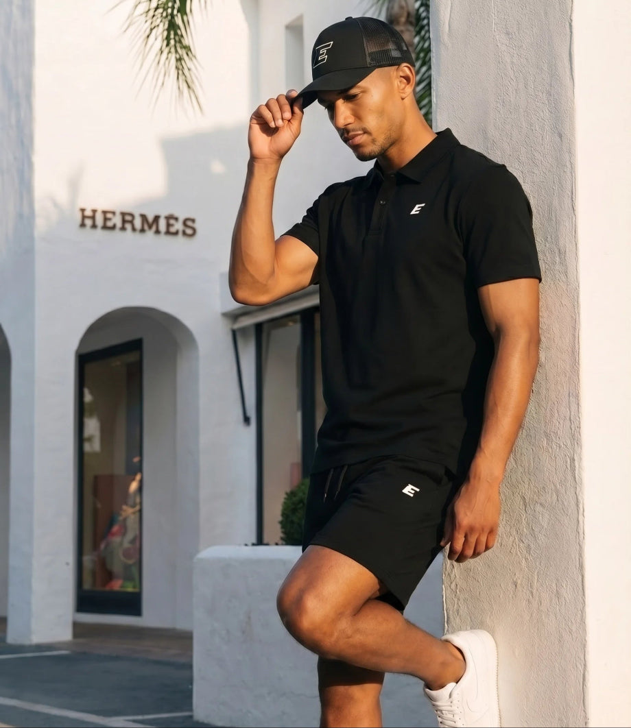 Man wearing a black polo shirt and shorts with a cap, standing in front of a Hermes store.
