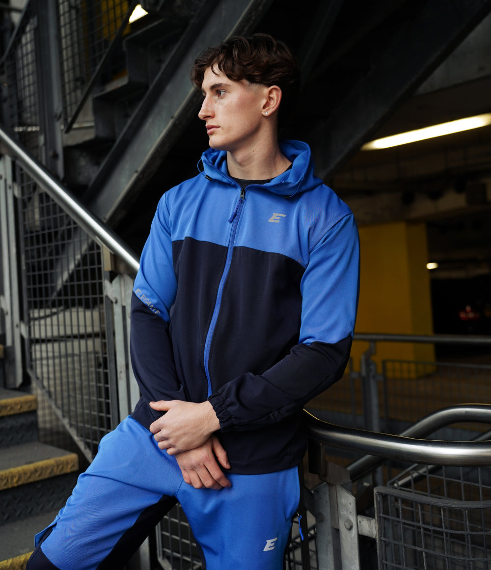 man wearing a blue and black tracksuit jacket with elegante brand logo, standing in an urban setting.