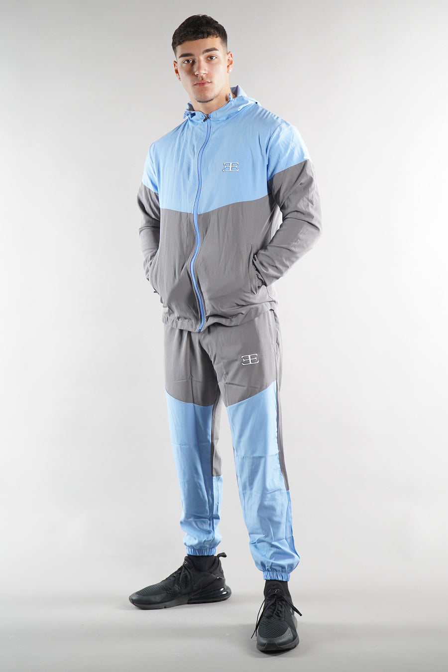 man wearing blue and grey elegante logo tracksuit jacket and joggers on white background