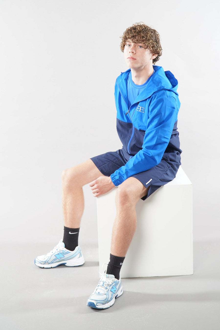 man wearing a blue hoodie and shorts sitting on a white chair against a light gray background