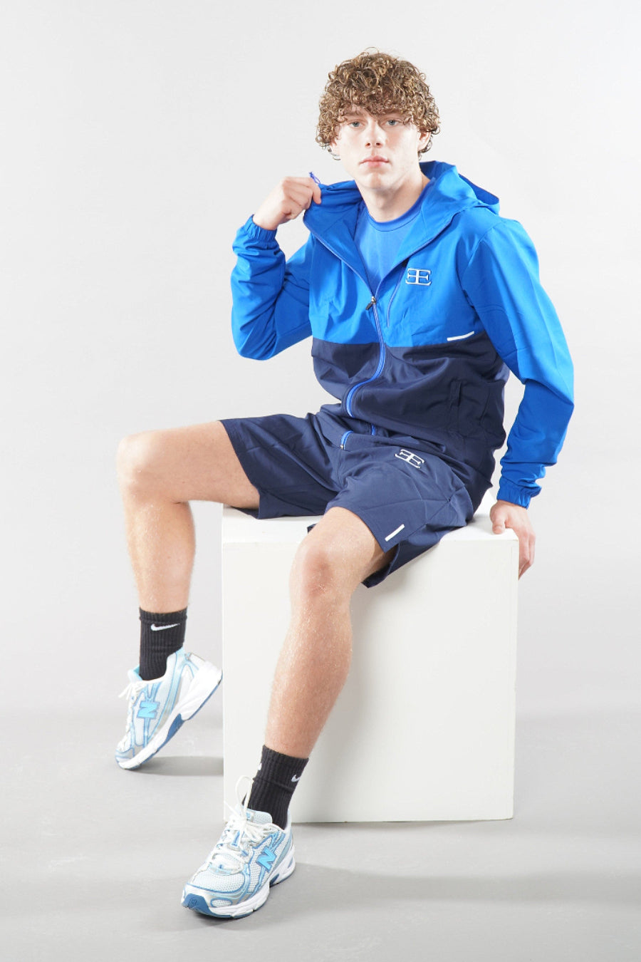 man wearing a blue athletic jacket and shorts sitting on a white cube against a light gray background