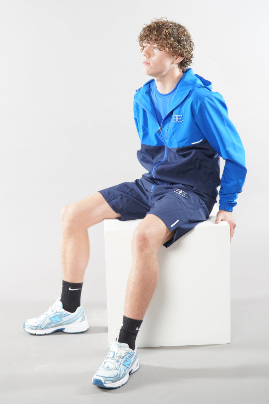 man wearing a blue and navy tracksuit sitting on a white cube against a light gray background