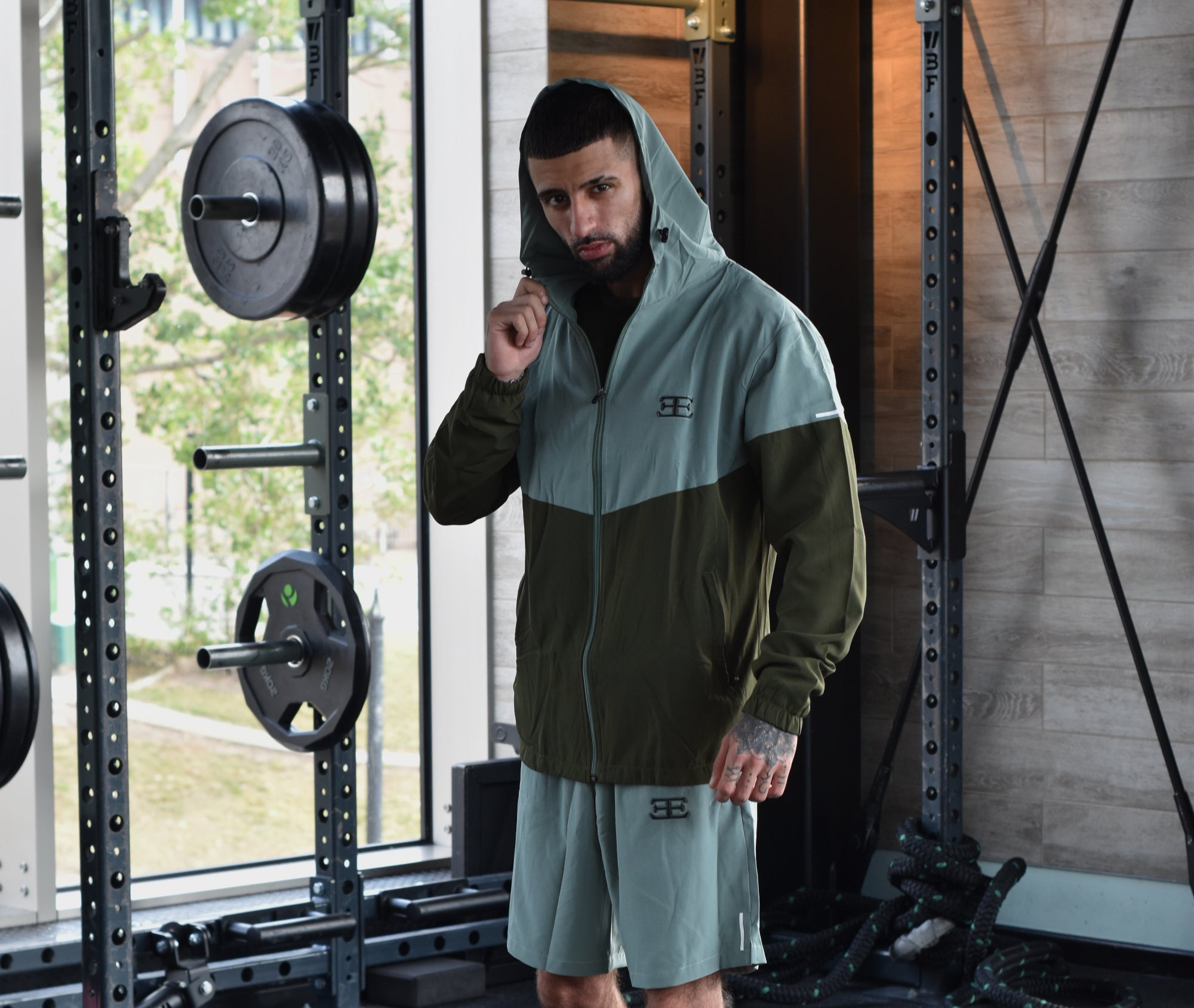 man wearing green gym clothing, shorts and jacket with hood up. with weights in the background.