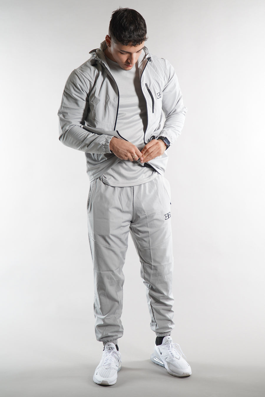 man wearing Elegante branded light grey Tracksuit jacket with hood and grey joggers on white background