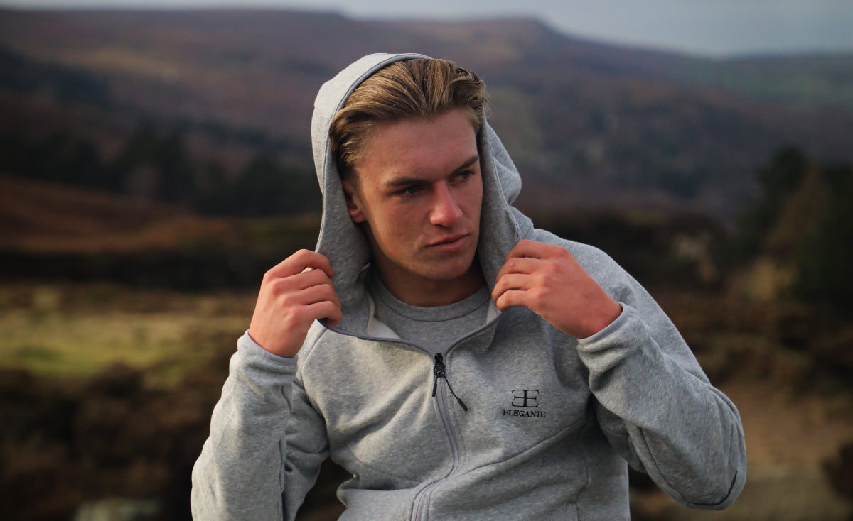 man with blonde hair wearing a grey zip up hoodie with hood up on countryside background
