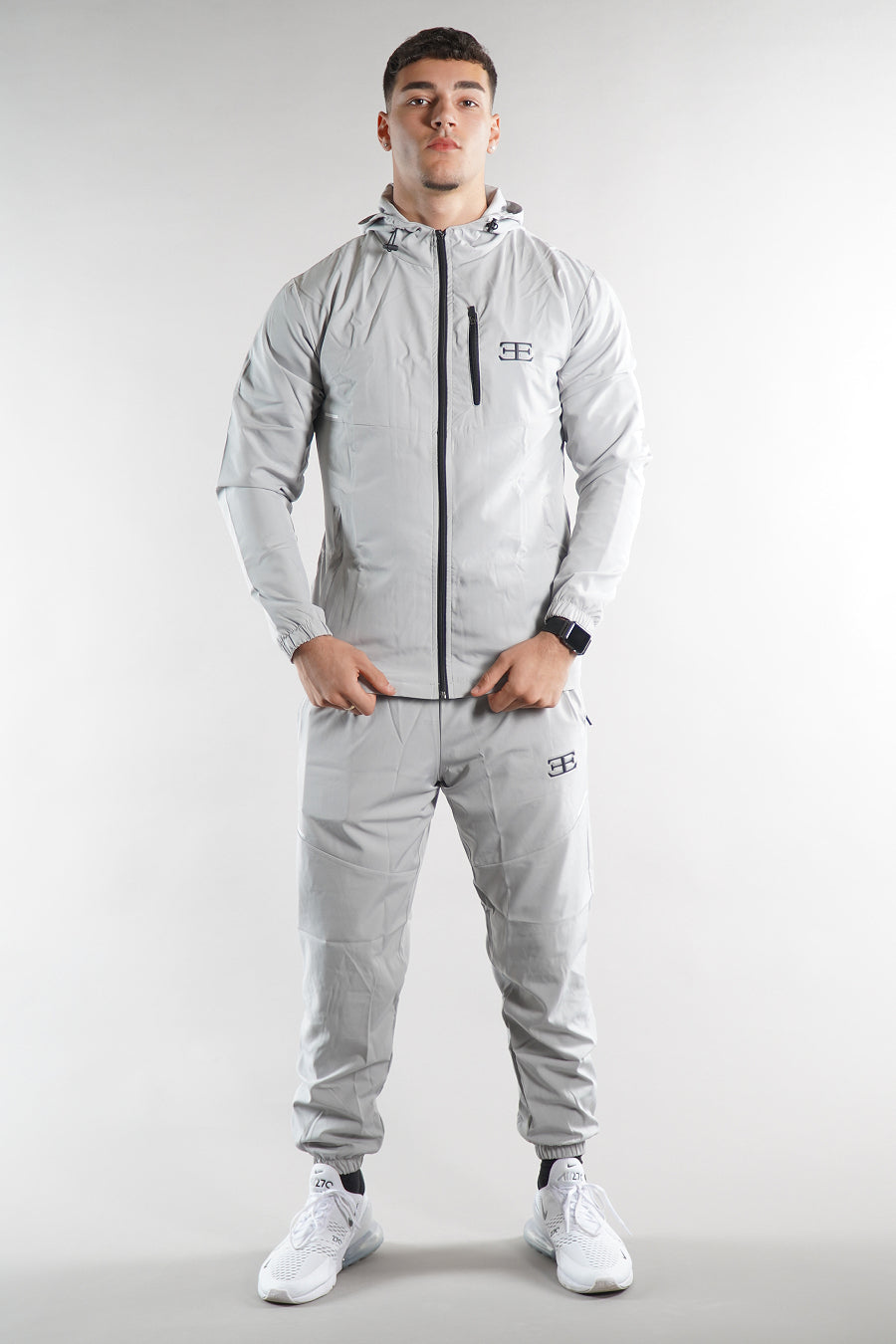 man wearing Elegante branded light grey Tracksuit jacket with hood and grey joggers on white background