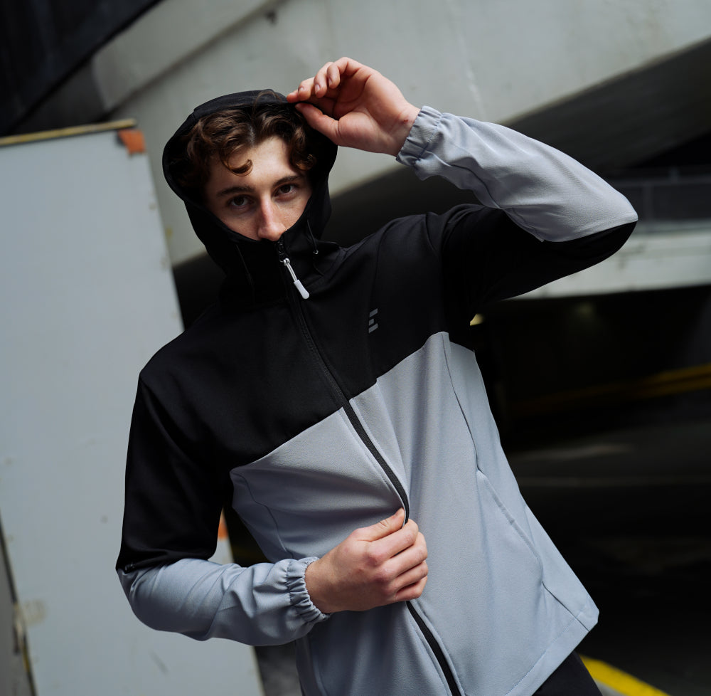 man wearing a black and gray tracksuit jacket with a hood, standing against an urban background.
